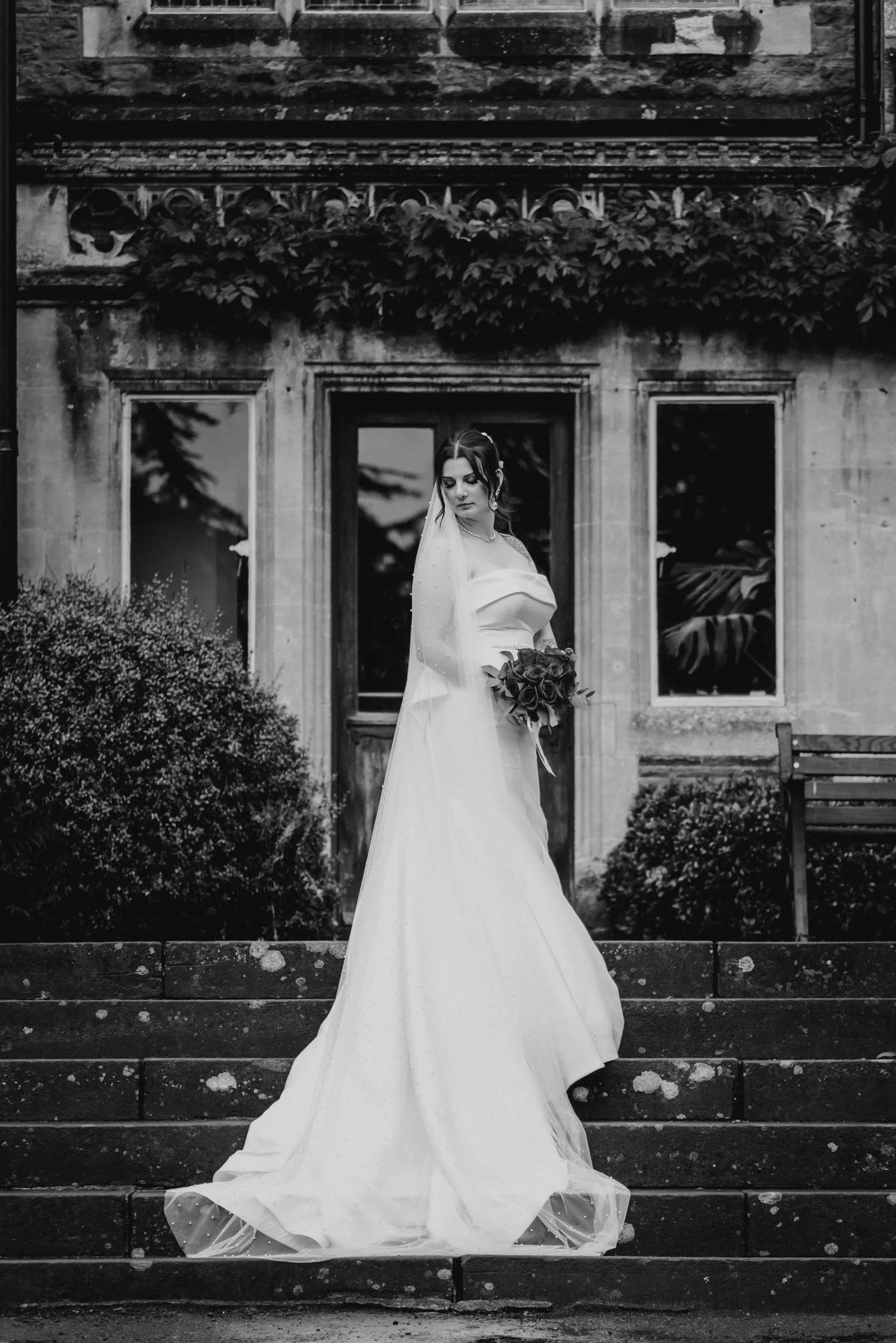 Black and white photo of a bride in a wedding dress holding a bouquet, standing on stone steps in front of an old building with overgrown plants and windows.