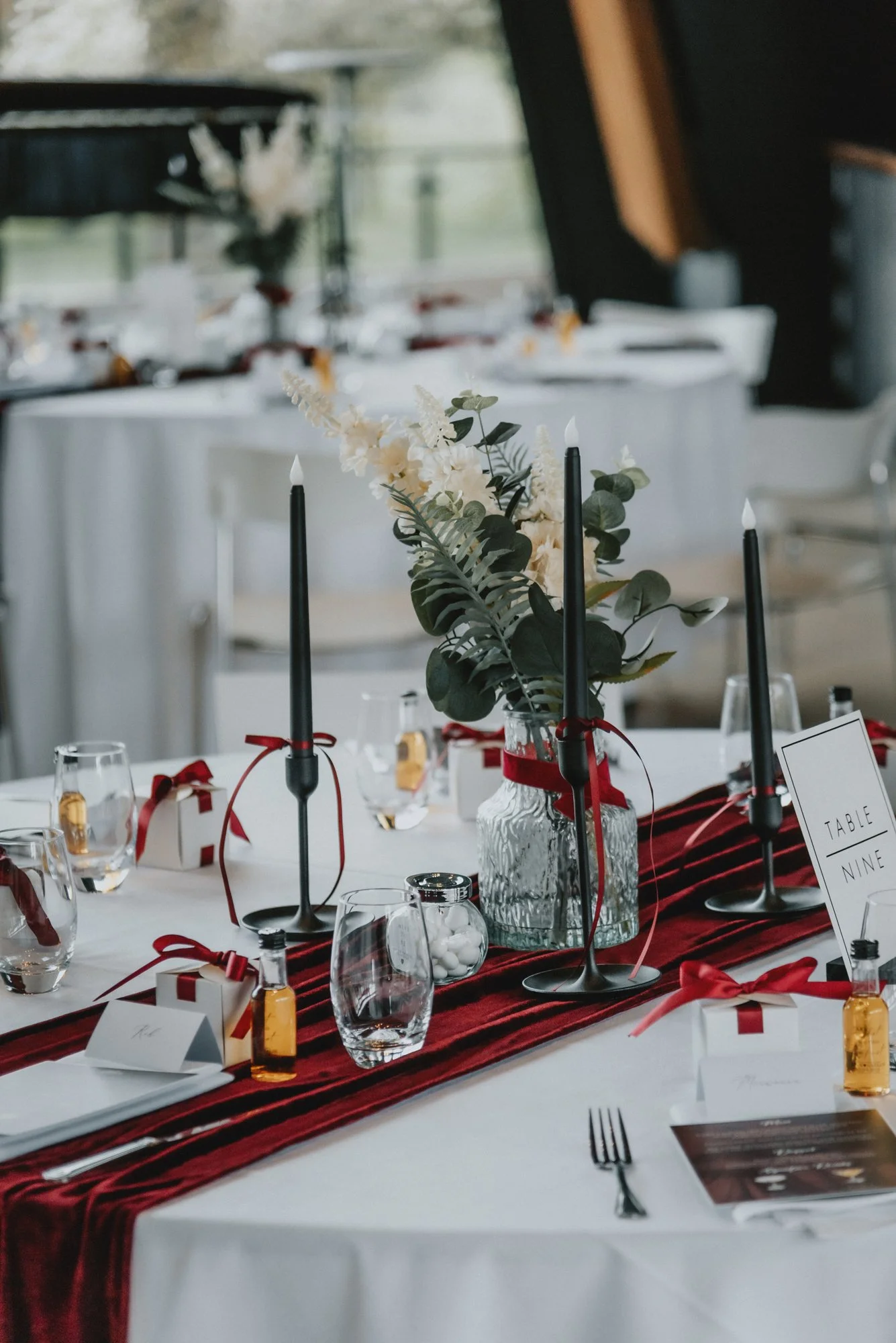 A decorated table for a formal event with a white tablecloth, a red striped table runner, and a floral centerpiece in a glass vase. The centerpiece includes white flowers and green foliage. The table has black candles, small gift boxes tied with red 