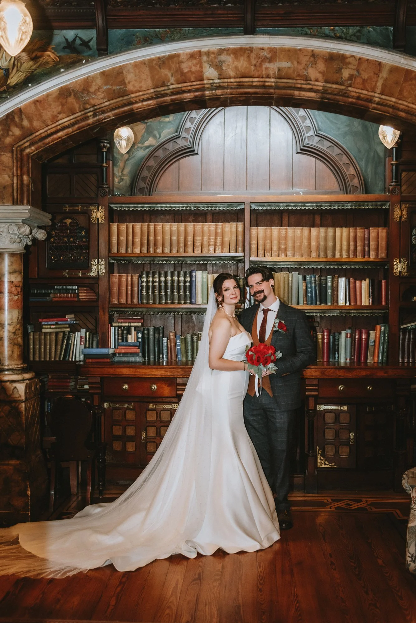 A newlywed couple stands in front of a vintage bookshelf filled with books, with a bride in a strapless white wedding gown with a long train holding a bouquet of red roses, and a groom in a dark plaid suit with a red tie and pocket square, smiling at