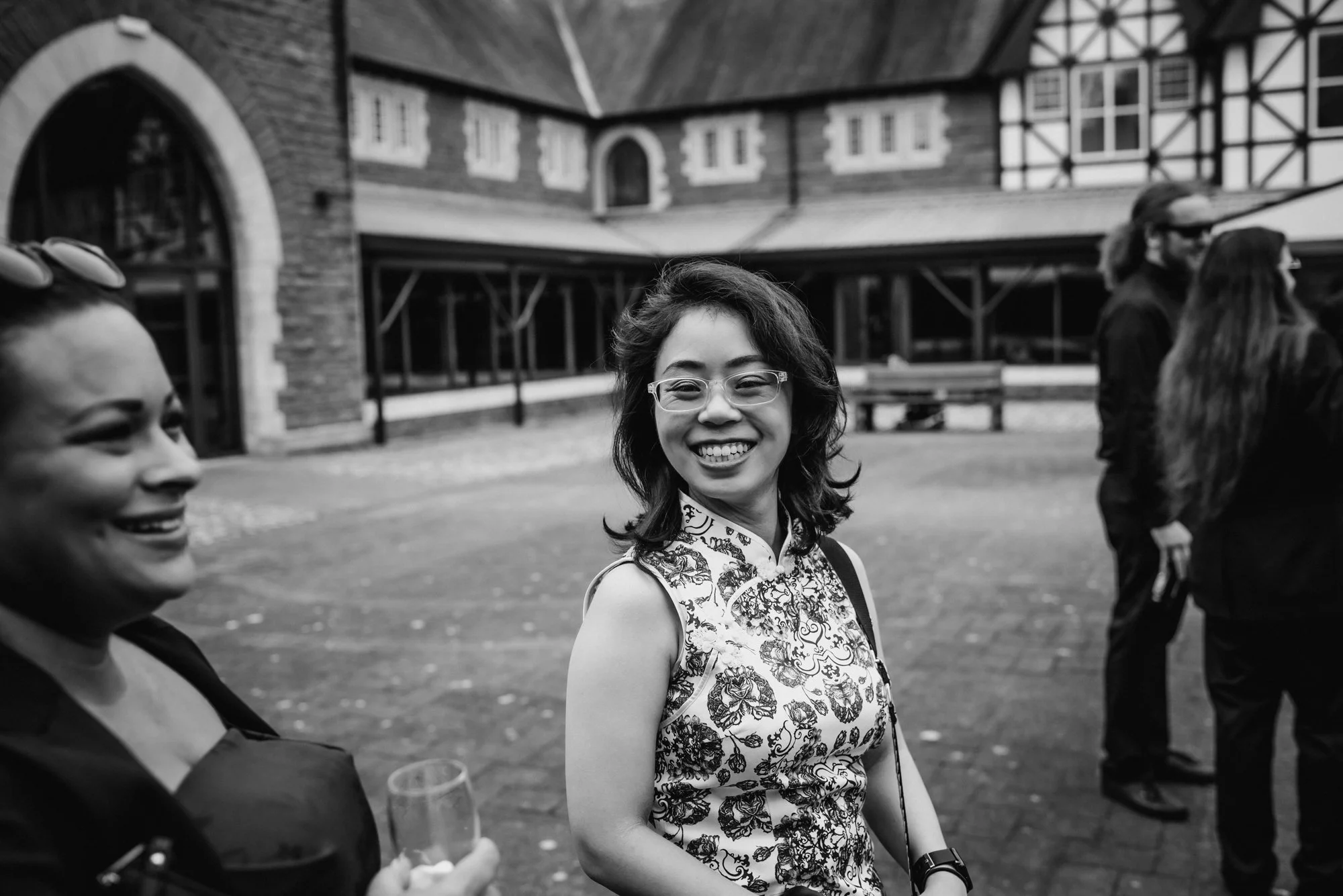 Smiling woman with glasses and floral top standing outdoors with other people nearby in front of a building.