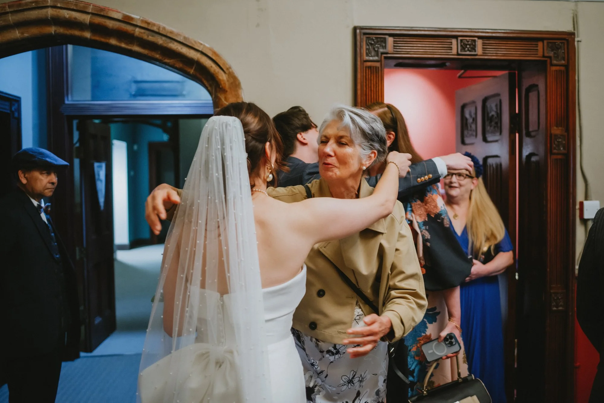 A bride in a white wedding dress and veil hugs an older woman, possibly her mother, in a warmly lit room. Other guests are visible in the background, hugging and smiling.