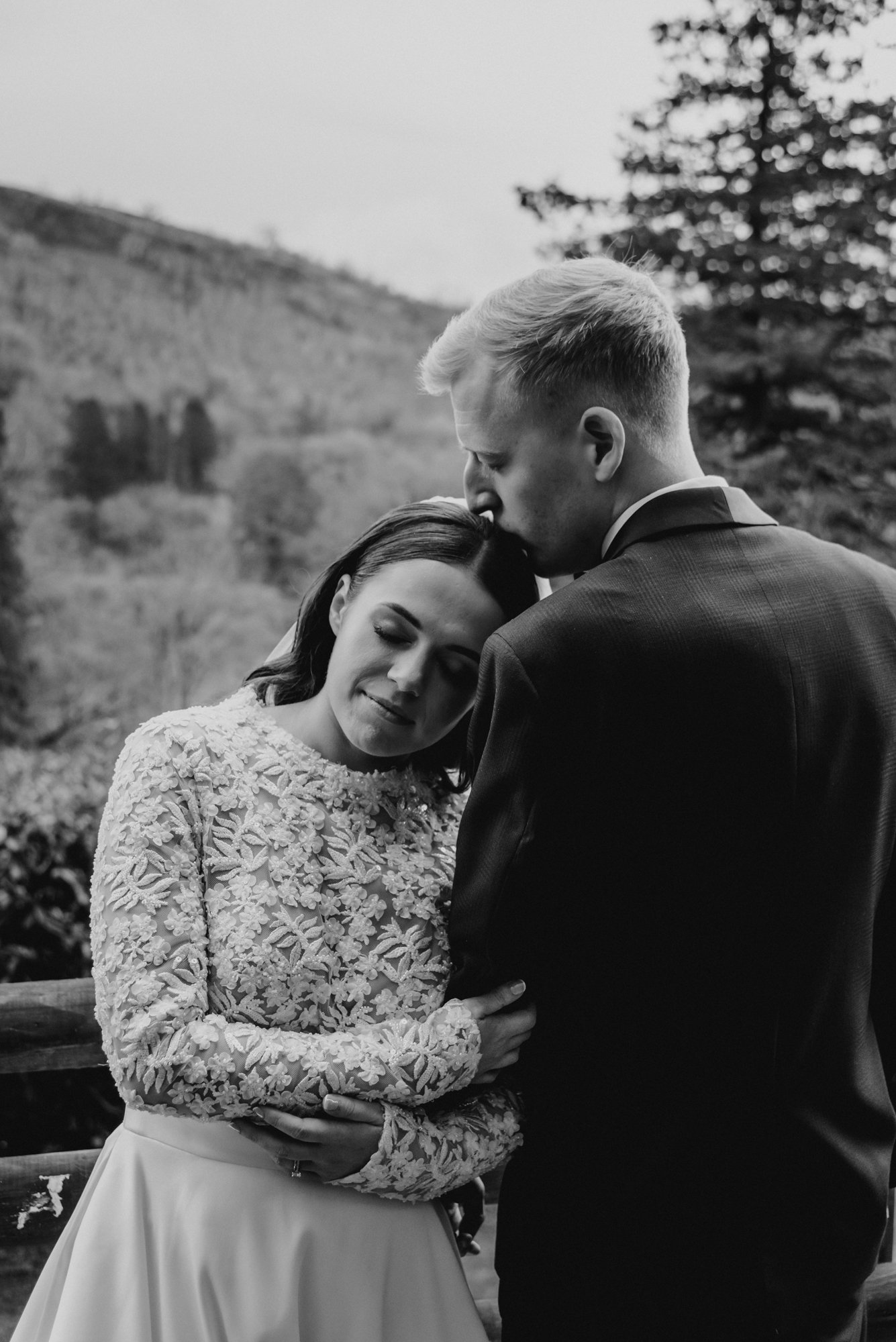 A black and white photograph of a bride and groom embracing outdoors. The bride has her eyes closed and is wearing a long-sleeved dress with floral lace detailing, while the groom is kissing her forehead.