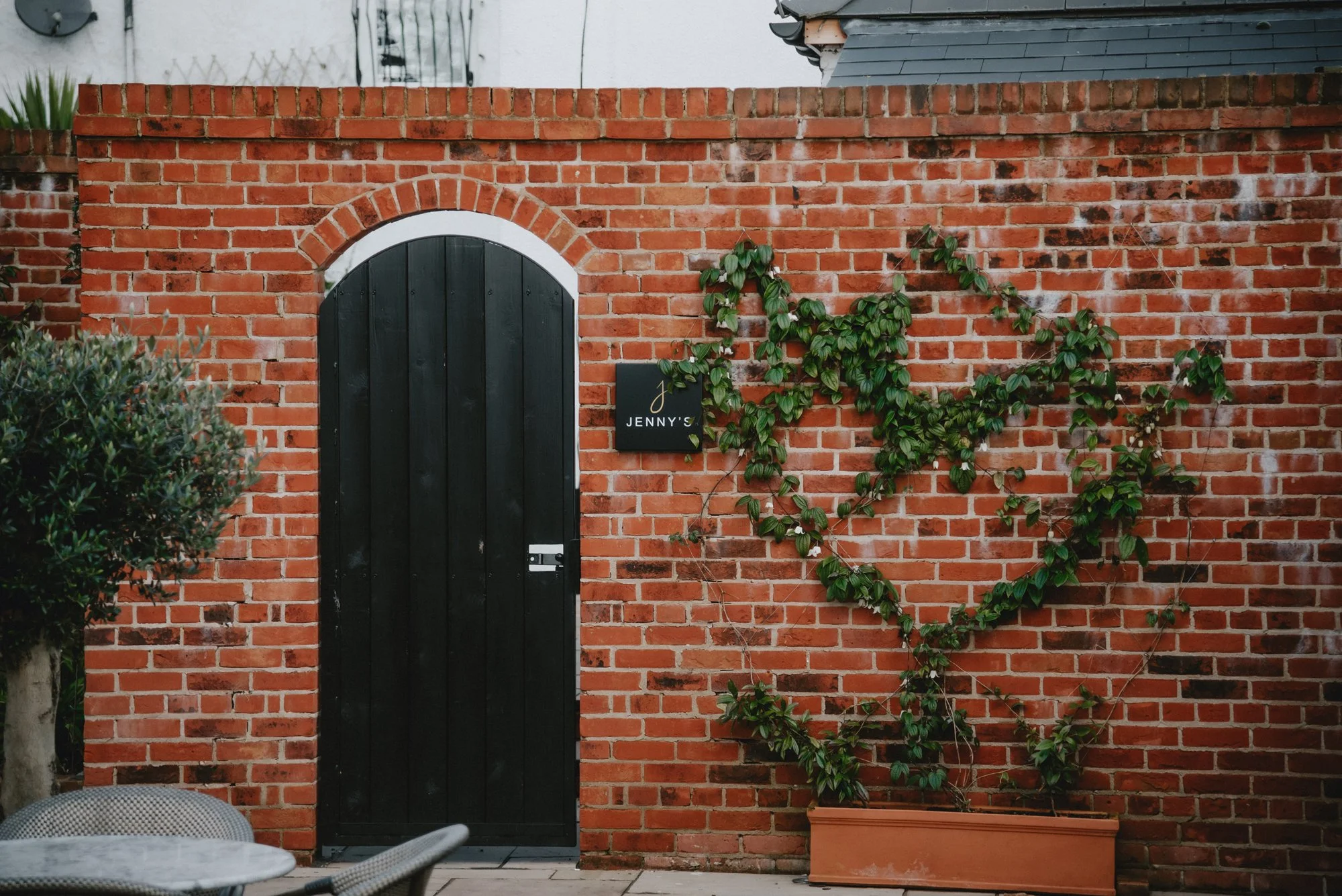 Brick wall with a black arched door, a small black sign reading 'JENNY'S,' and a heart-shaped vine decoration, with outdoor furniture and a tree nearby.