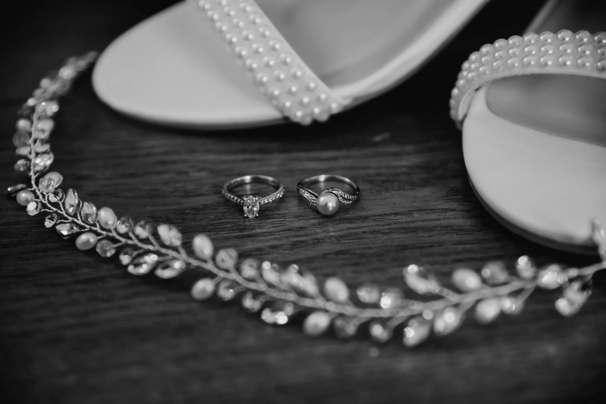 Black and white photo of a pearl ring, a diamond ring, a pearl bracelet, pair of white high-heeled shoes with pearl embellishments, and a pearl hairband on a wooden surface.