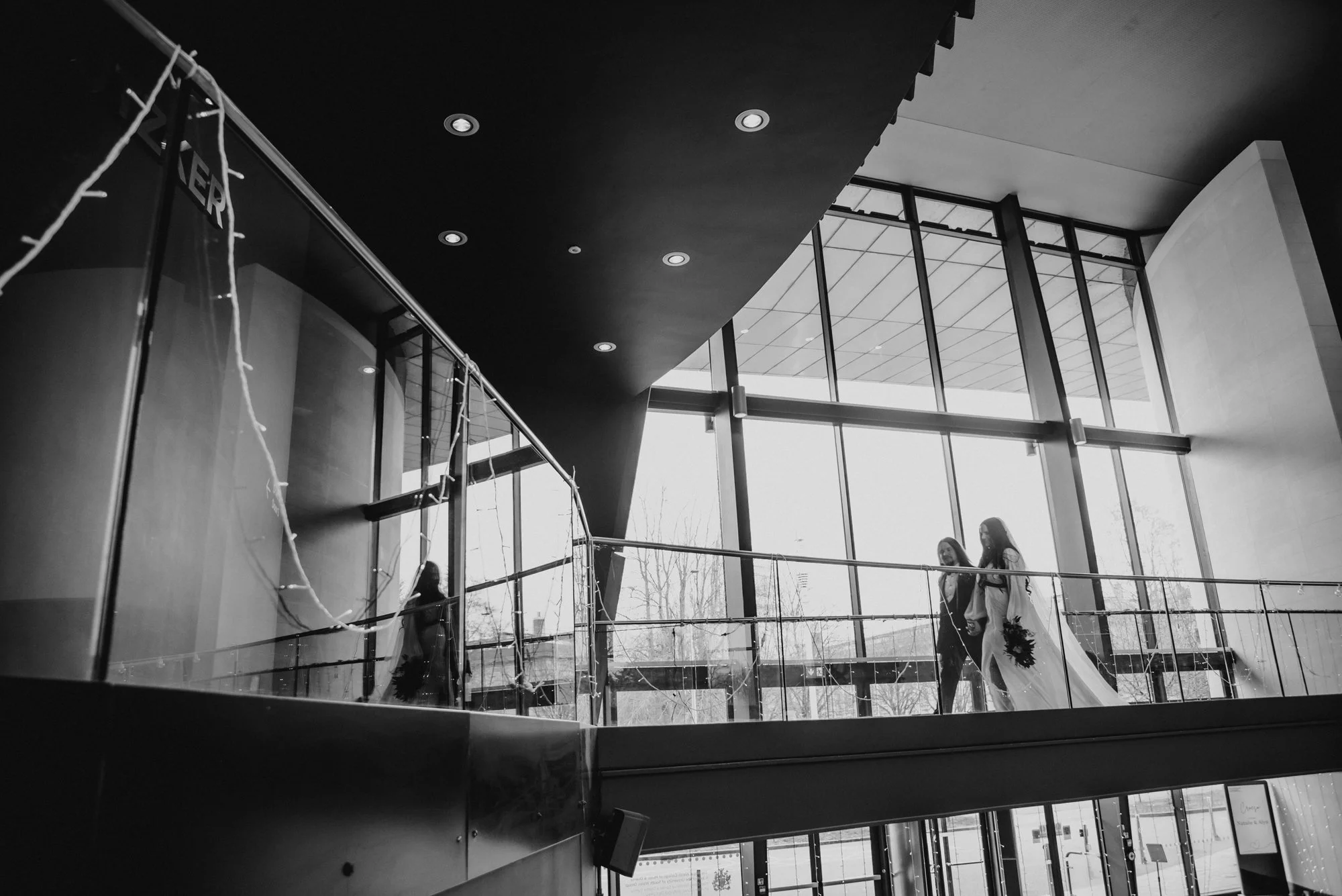 Two women, one in a dark outfit and the other in a white wedding dress, walking together inside a modern building with large glass windows and high ceilings.