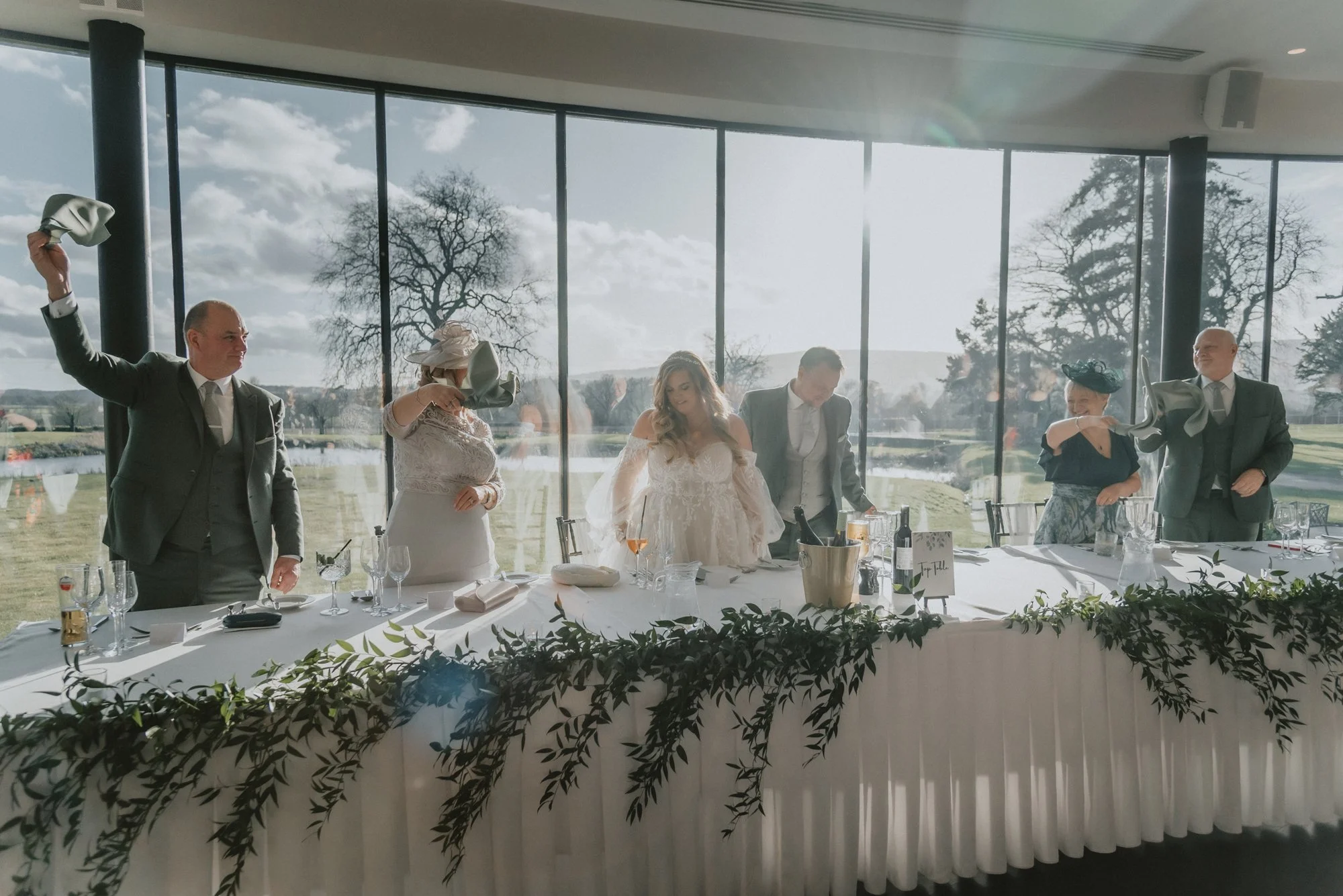 A wedding reception with a bride and five guests tossing napkins in the air at a long, decorated table. The reception is in a glass-walled venue with a scenic outdoor view, including leafless trees, a lake, and a partly cloudy sky.