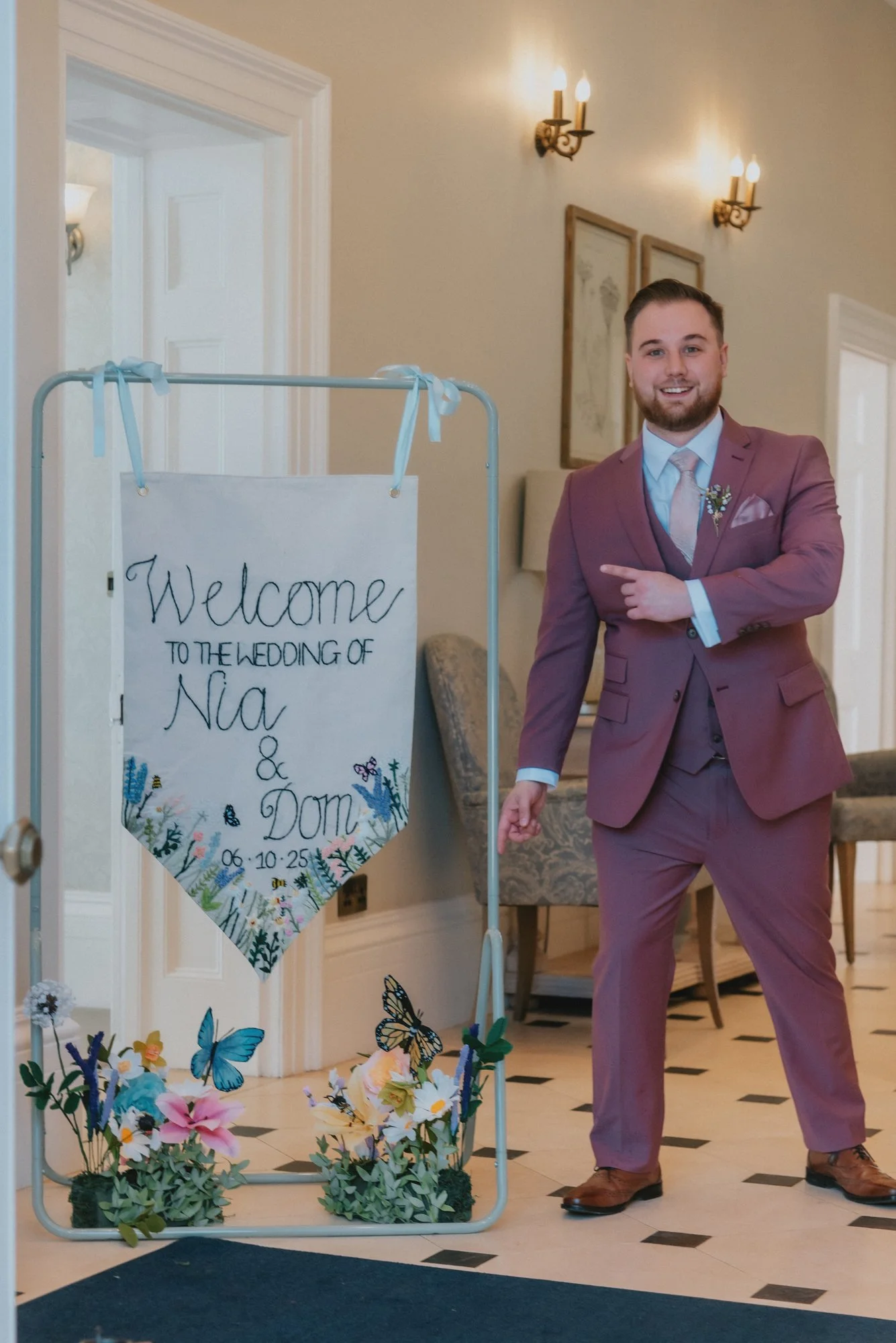 A man in a maroon suit pointing to a welcome sign for a wedding, decorated with flowers and butterflies.