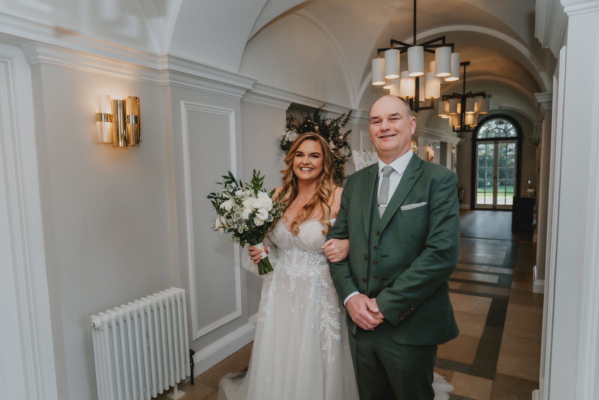 A bride in a white wedding dress holding a bouquet of white flowers and greenery, standing next to a man in a green suit inside a well-lit elegant building.