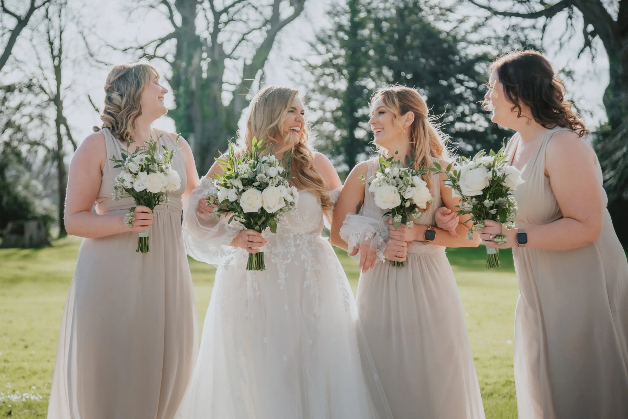 A bride and four bridesmaids standing outdoors in a park, holding bouquets of white roses and greenery, smiling and enjoying each other's company.