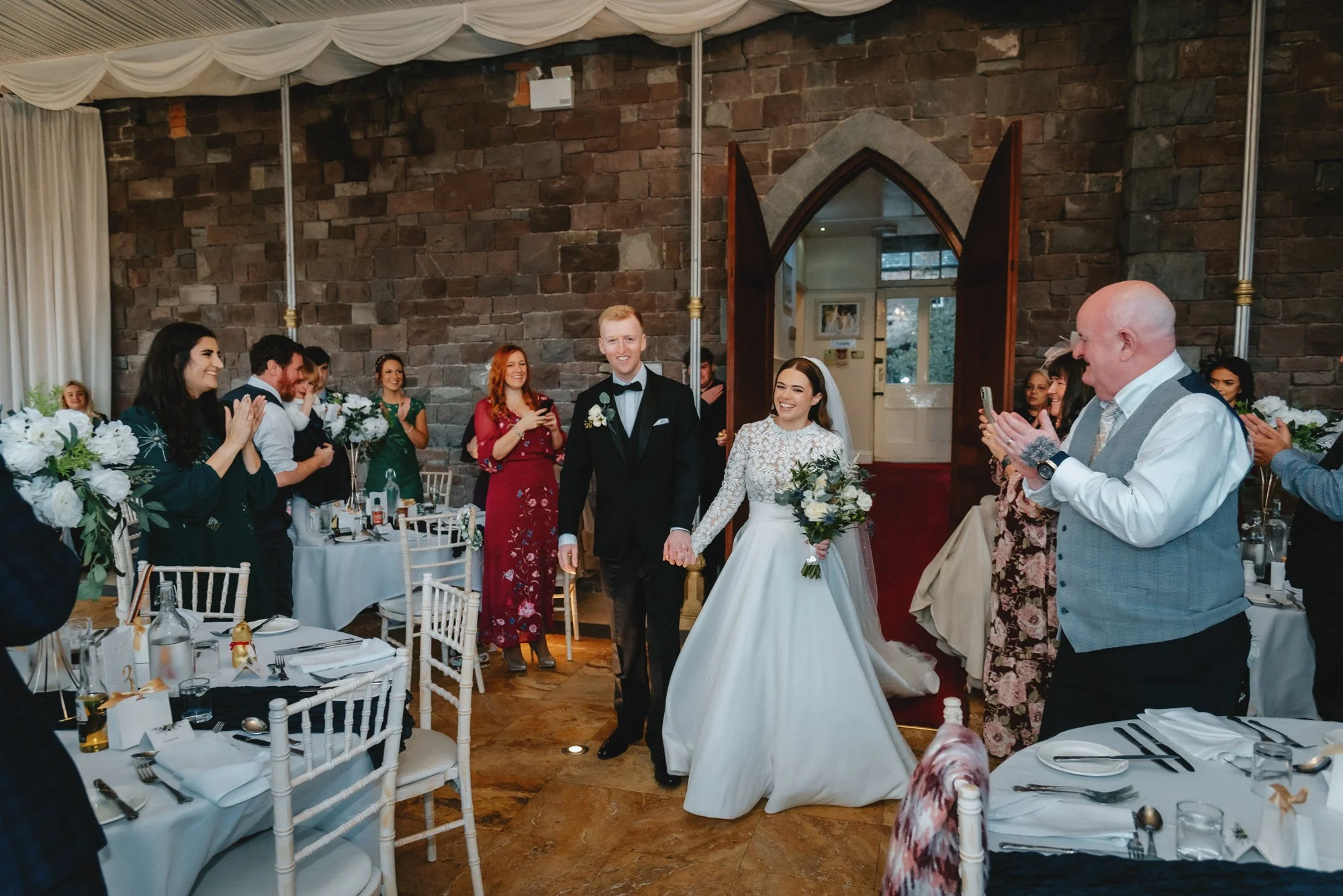 Newlywed couple holding hands walking into their wedding reception, guests clapping and smiling, decorated tables with floral arrangements, warmly lit indoor venue with stone walls and arched doorway.