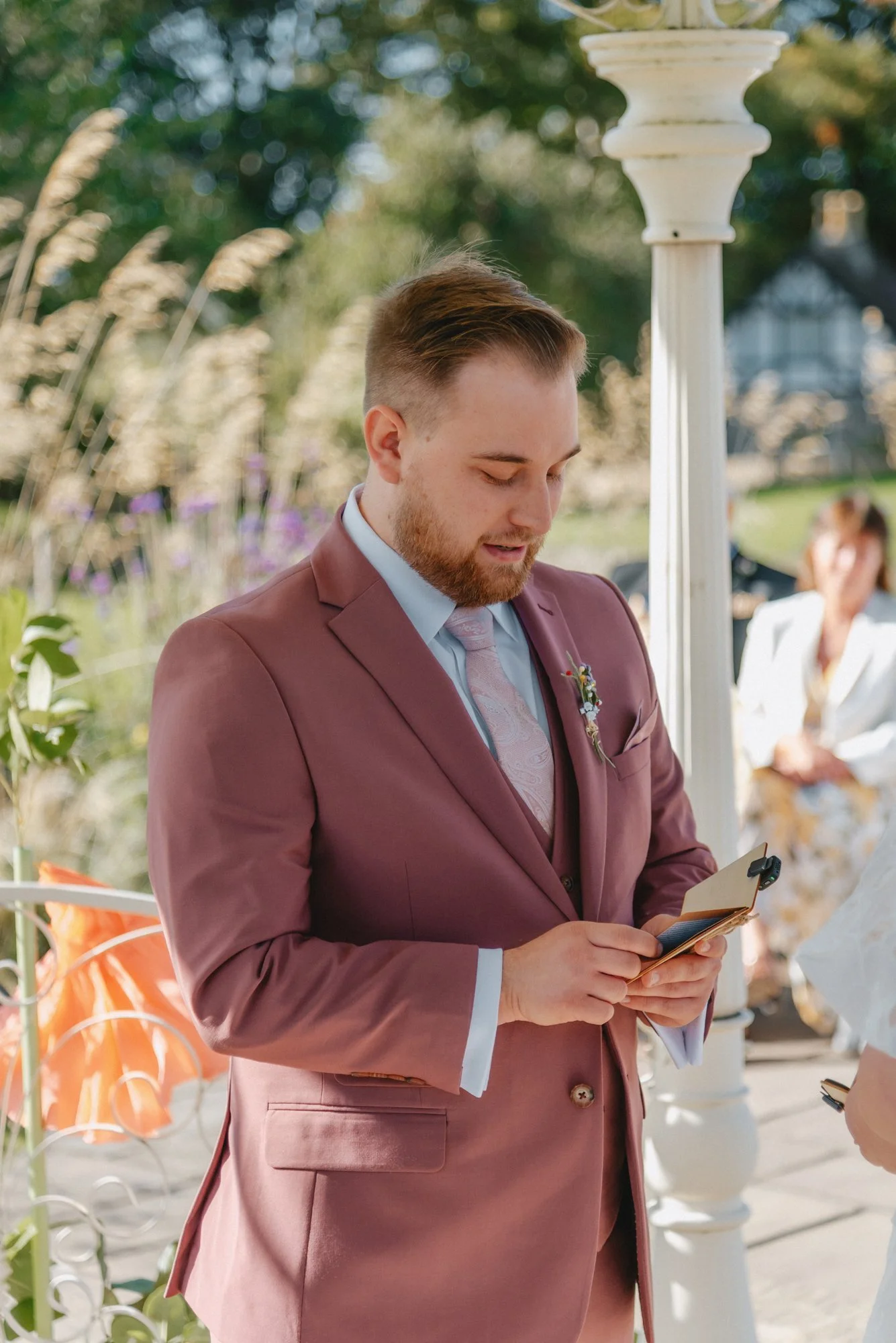 Groom in a mauve suit standing outdoors, reading a small card at a wedding ceremony.