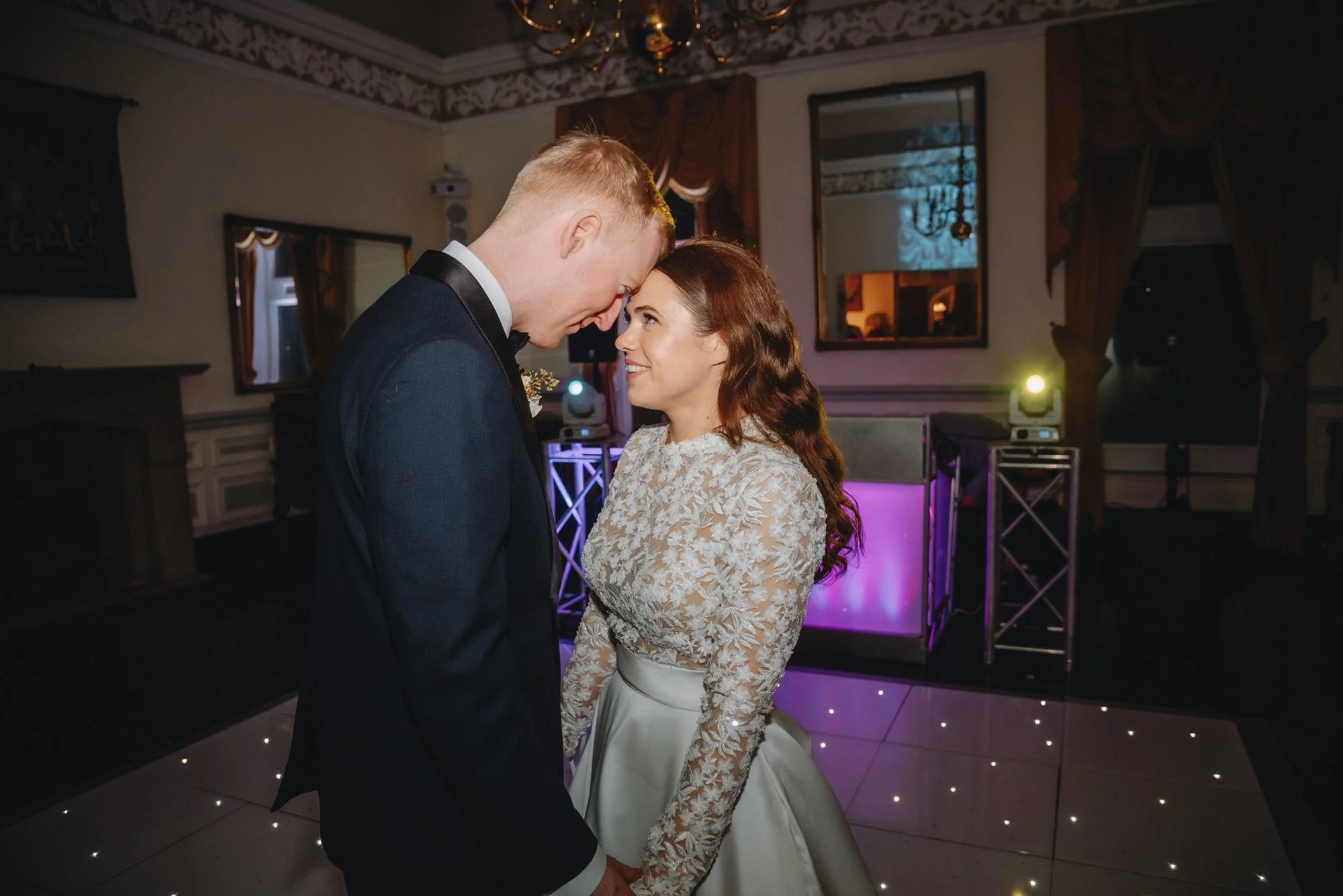 A bride and groom smiling and holding hands during their wedding reception dance in a decorated ballroom with lighting and mirrors.