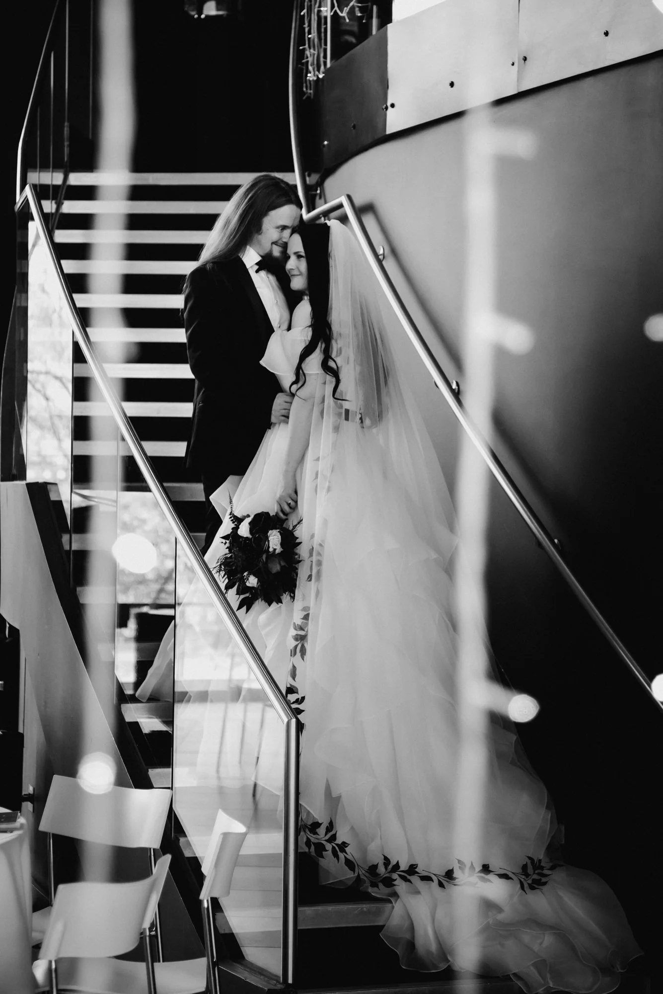A bride and groom on a staircase, sharing an intimate moment during their wedding, with the bride holding a bouquet of flowers, both smiling.