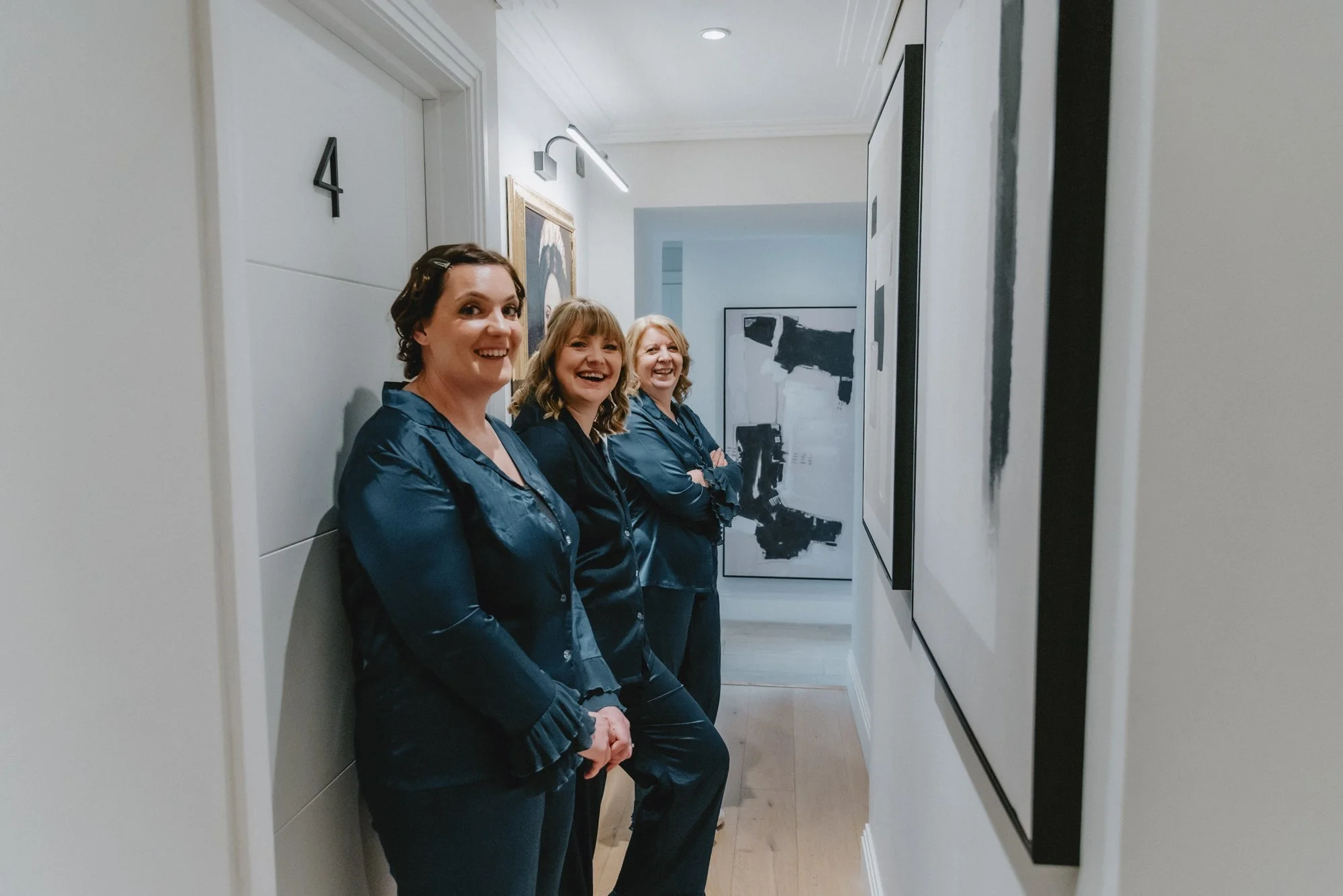 Three women in navy blue pajamas standing in a hallway, smiling and looking at the camera.