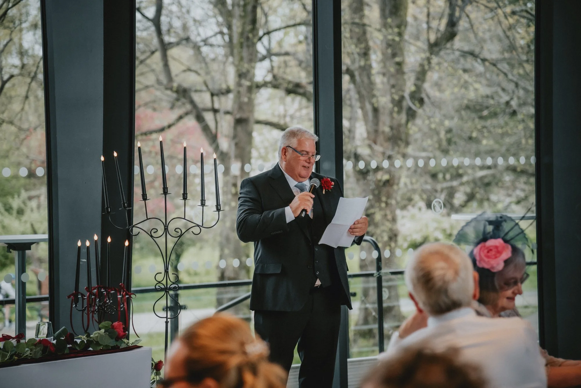An elderly man in a black suit with a red flower lapel pin is giving a speech with a microphone at a wedding reception. Behind him is a large window showing trees, and to his left is a tall black candelabrum with lit candles. Guests are seated and li