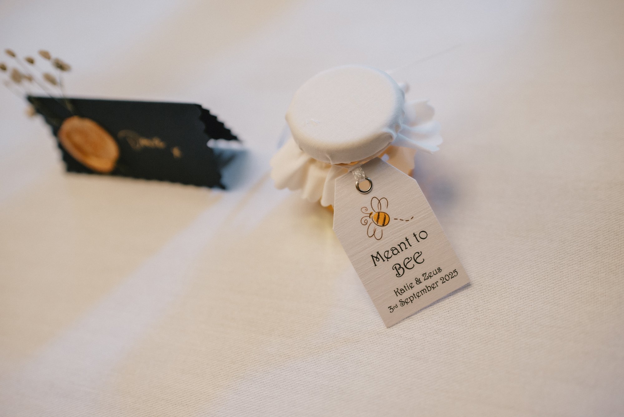 A small wedding favor with a white fabric top, a bee illustration on the tag, and text that reads 'MANT TO BEE, Katie & Zeus, 3rd September 2025' on a white tablecloth.