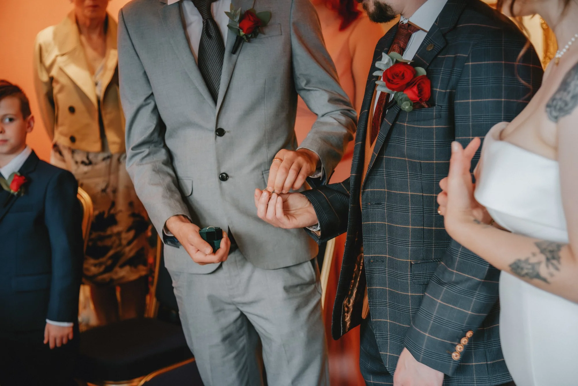 A man in a gray suit is placing a ring on another man’s finger, who is wearing a dark plaid suit with a red rose boutonniere, at a wedding ceremony. A woman with tattoos in a white dress is standing nearby, and a young boy in a suit sits watching. Ad