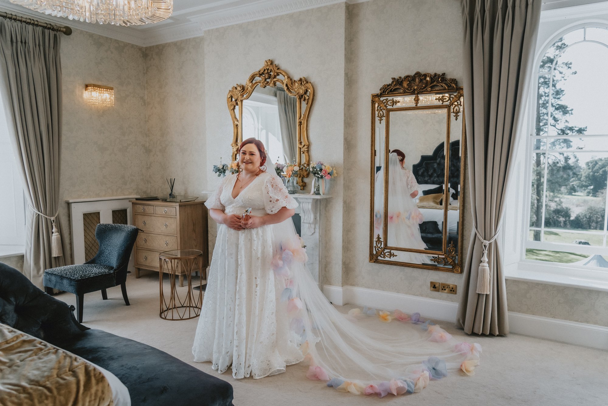 A woman in a white lace wedding dress standing in a beautifully decorated room, smiling with a long veil made of colorful flower petals flowing behind her as she holds a small object in her hands, with ornate gold-framed mirrors on the wall and large