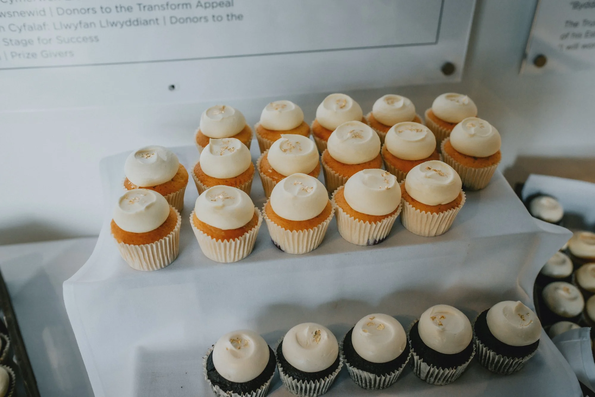 A display of cupcakes with white frosting, some topped with gold leaf, arranged on a tiered stand at an event or bake sale.
