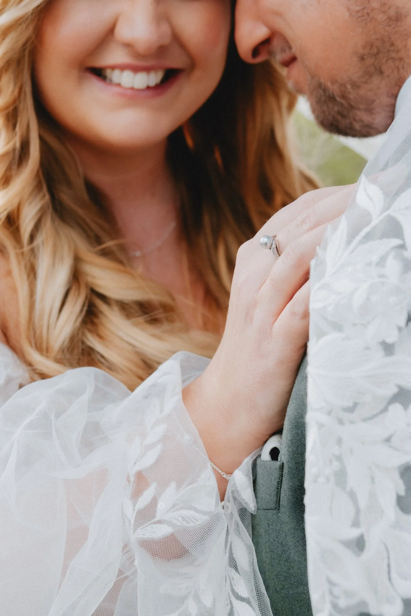 Close-up of a smiling woman with blonde hair and a man, showing their faces and an engagement ring on her finger, with the woman gently resting her hand on the man's chest.