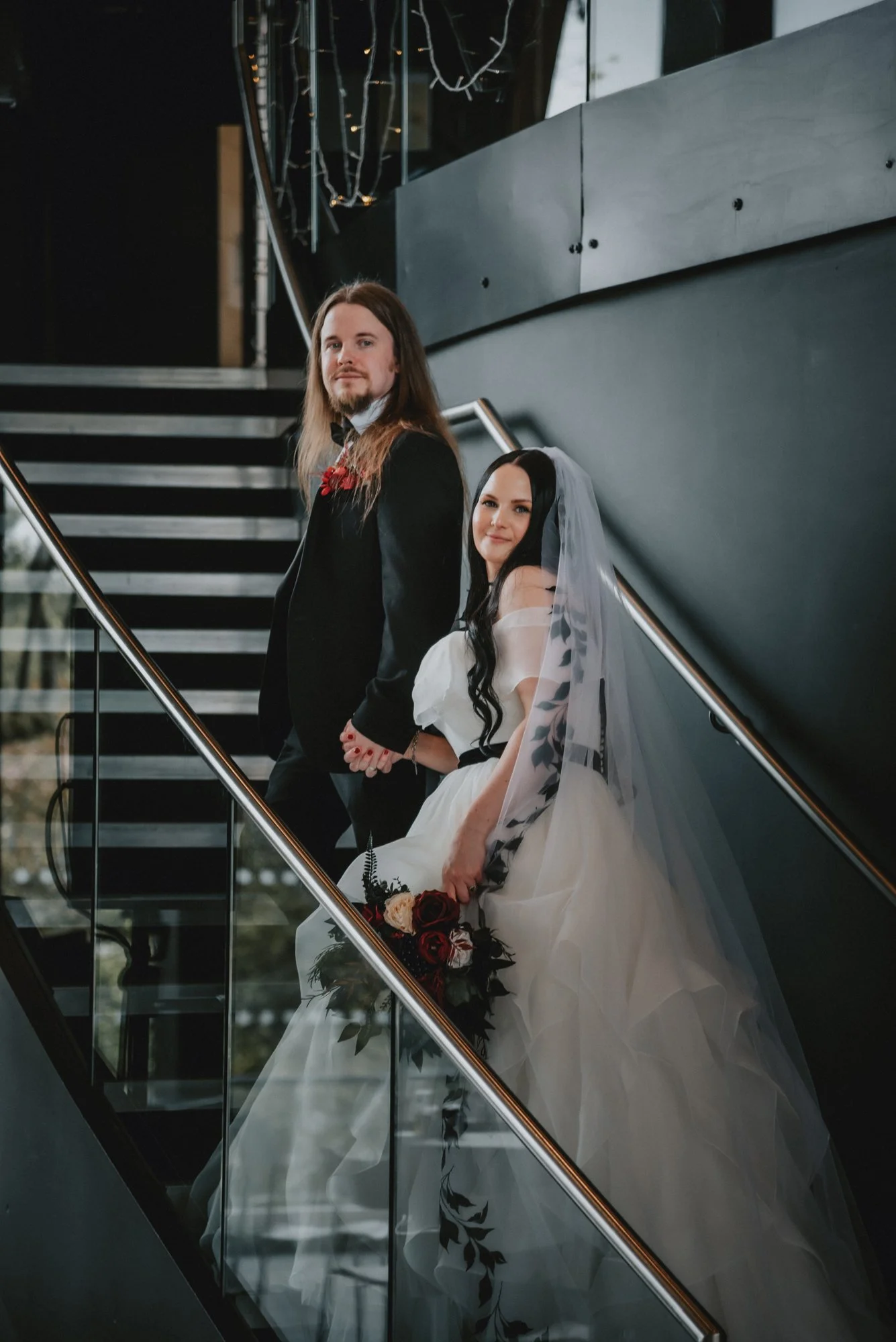 A bride and groom standing on a staircase at their wedding, holding hands and looking at the camera, with the bride in a white dress and veil holding a bouquet of red and white flowers, and the groom in a black tuxedo with a bow tie.