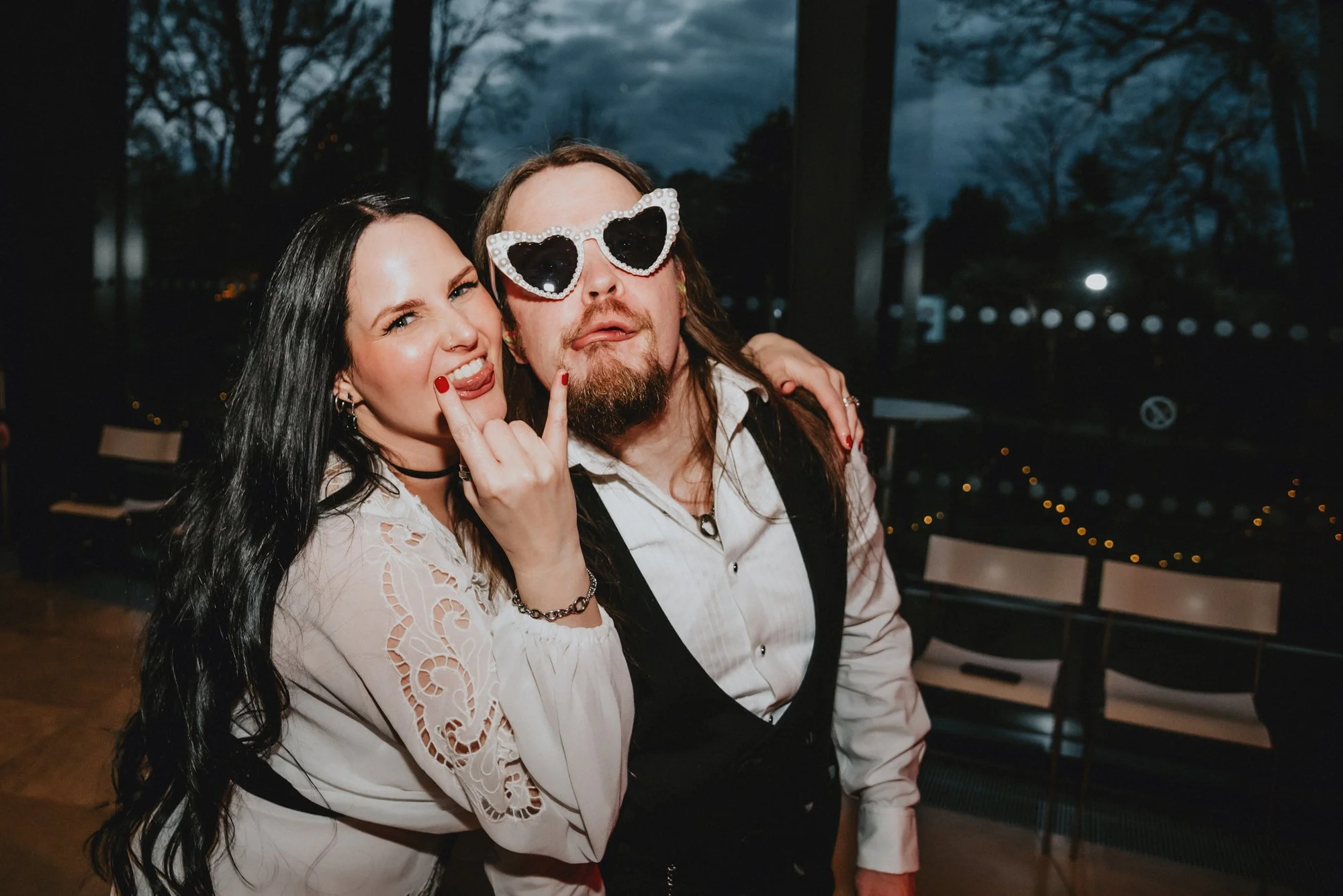 A woman with black hair and a man with long hair and a beard are posing together, making rock-on gestures and sticking out their tongues. The woman is wearing a white blouse with lace details, a choker, and red nail polish. The man is wearing heart-s