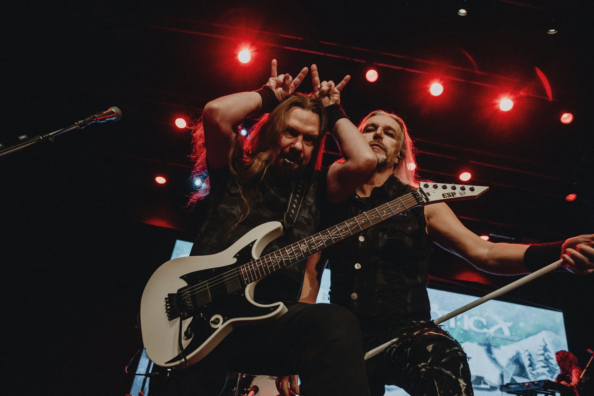 Two rock musicians on stage, one holding a white electric guitar and making a playful gesture with his hands on his head, the other holding a microphone stand, with red stage lights overhead and a large screen in the background.
