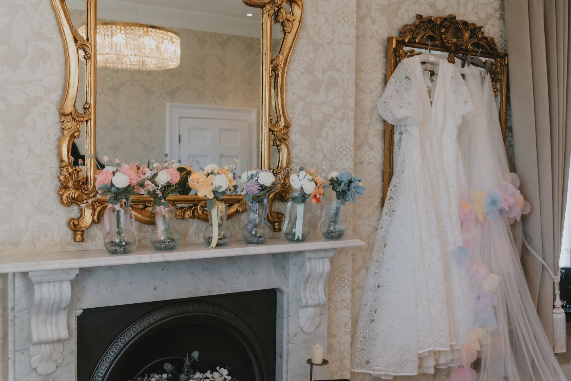 A decorated room with a large ornate gold-framed mirror above a white marble fireplace mantel. On the mantel, five glass vases hold pastel-colored artificial flower arrangements. To the right, a gold-framed dress with lace details and colorful flower