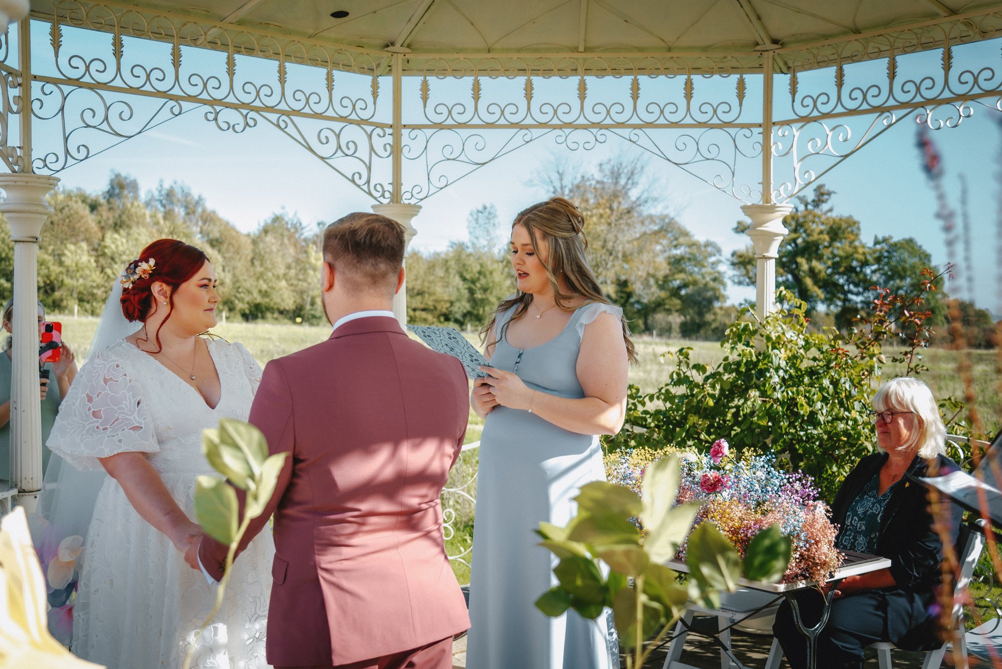 A couple getting married under a gazebo with a woman officiating, outdoors on a sunny day, surrounded by greenery and flowers, with a woman playing a keyboard in the background.