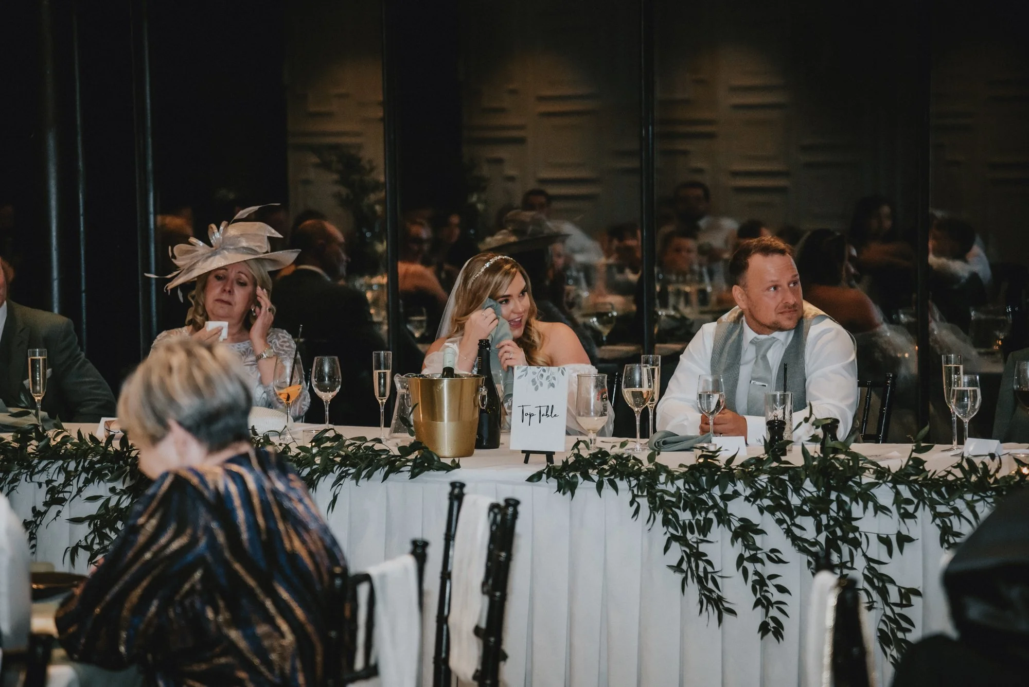 People sitting at a long banquet table during a wedding reception, with a woman in a white dress and a veil wiping her eye, a woman in a large hat talking on the phone, and a man in a white shirt and gray vest looking to the side.