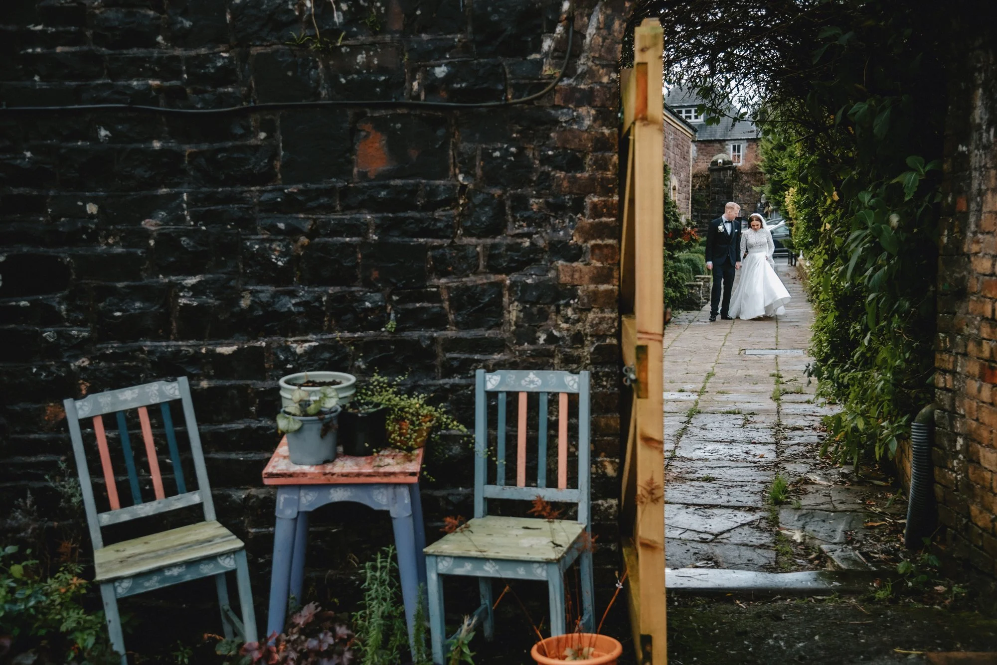 A bride and groom walking outdoors on a stone pathway, framed by greenery and brick walls, after a wedding ceremony.