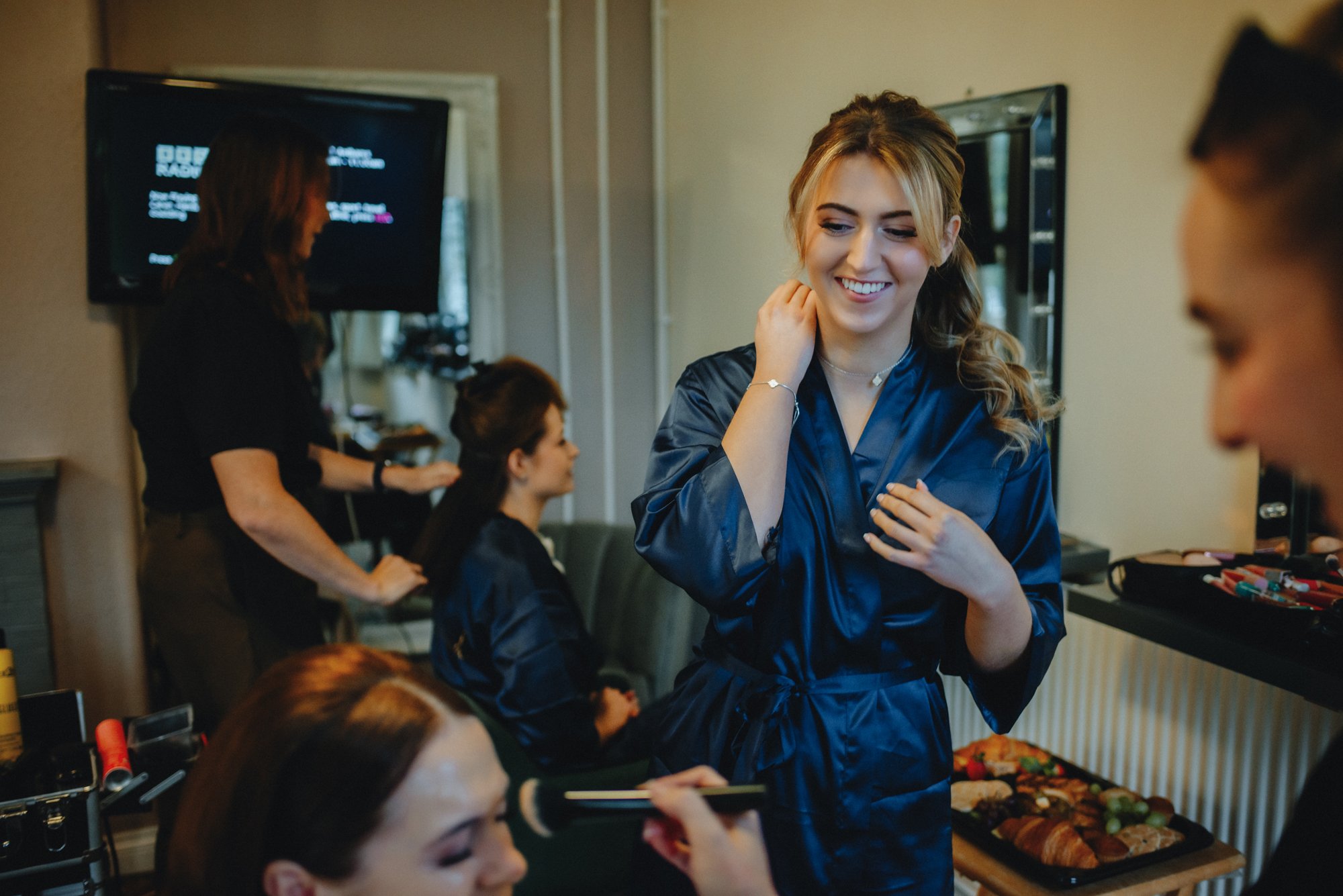 Women getting ready, with one woman at the forefront smiling, in a room with mirrors and beauty products.