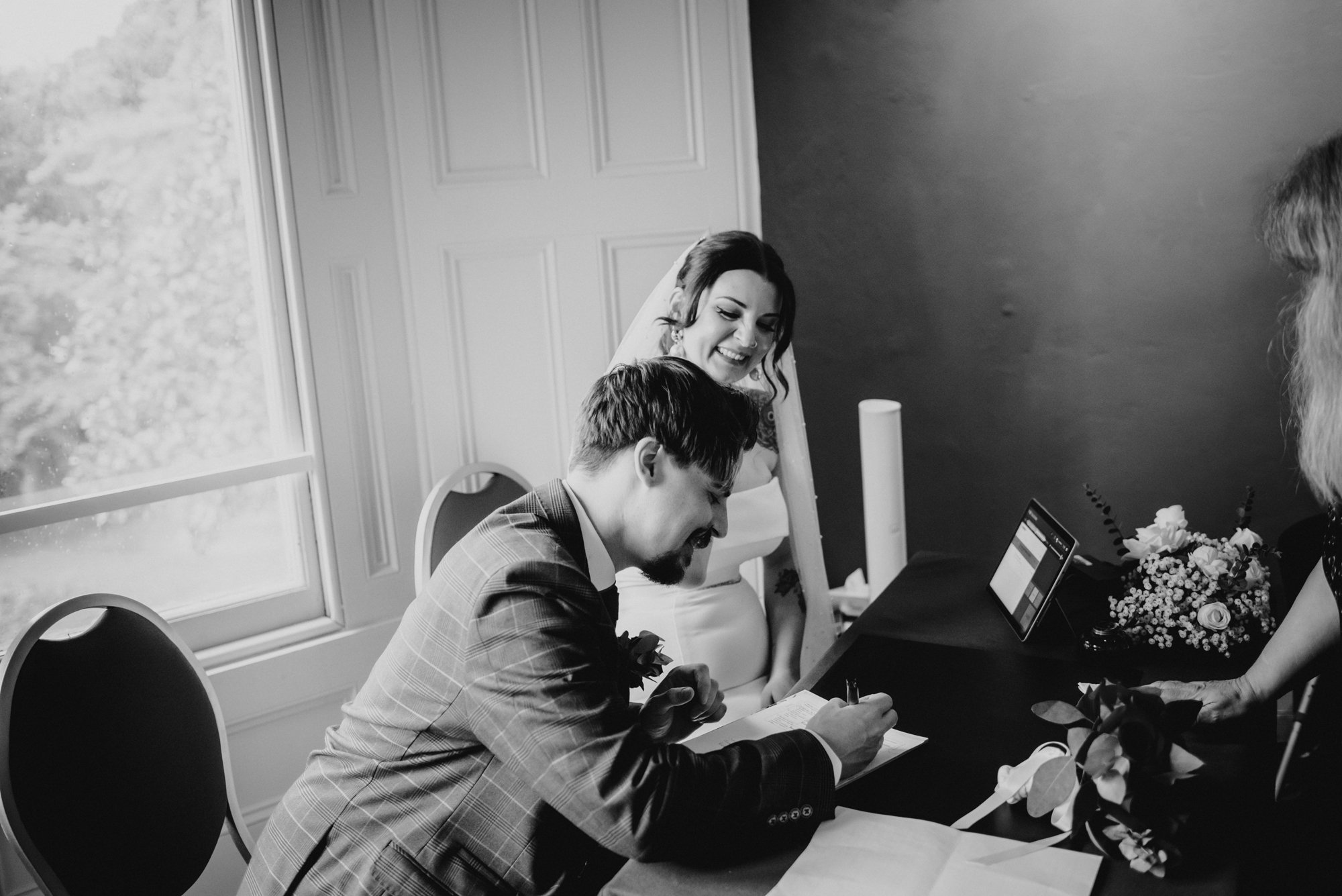 A bride and groom signing marriage documents during their wedding ceremony in a room near a window, with a floral bouquet and an officiant present.