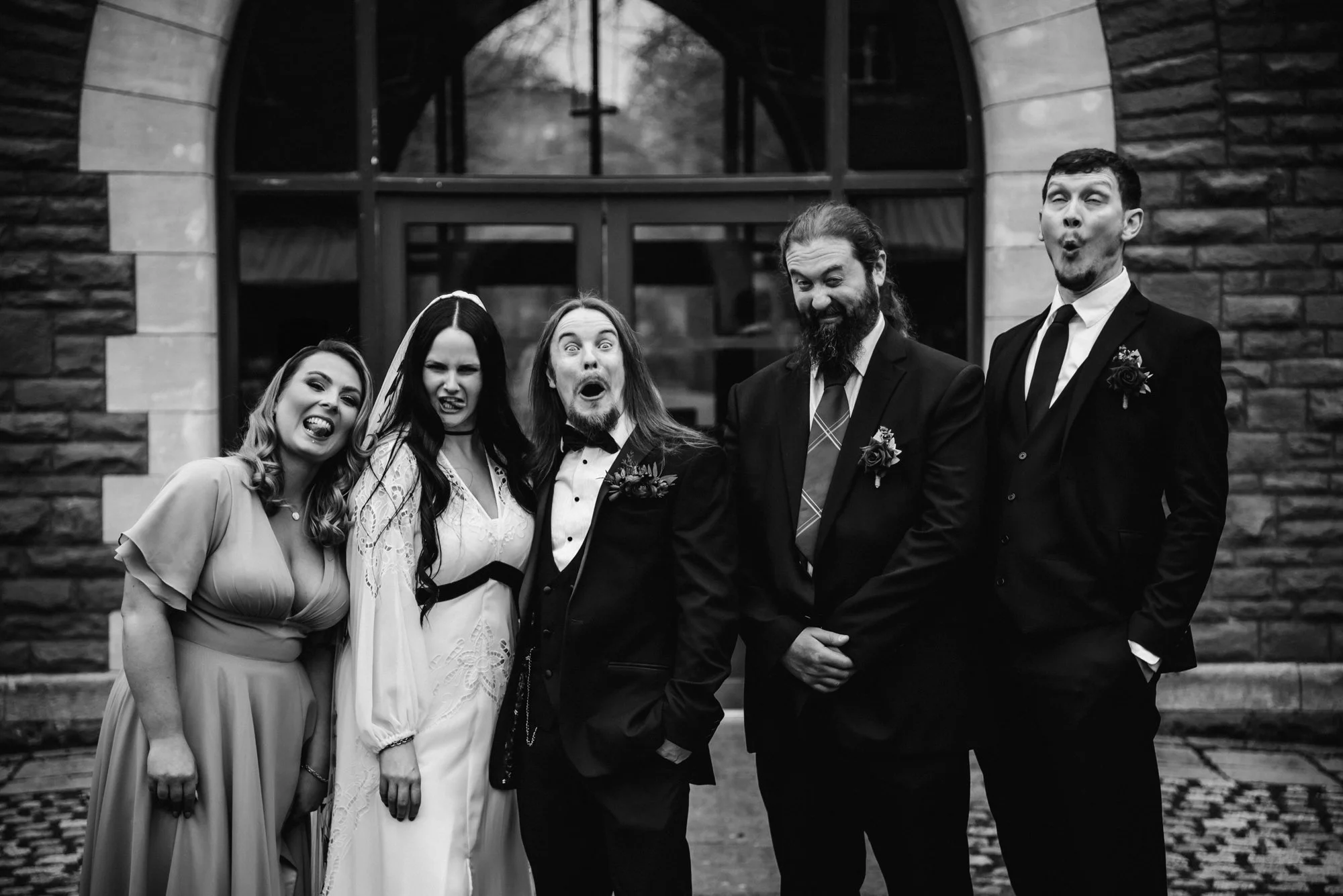 A group of five people dressed in formal wedding attire posing in front of a stone building with arched window. They are making playful, silly faces.