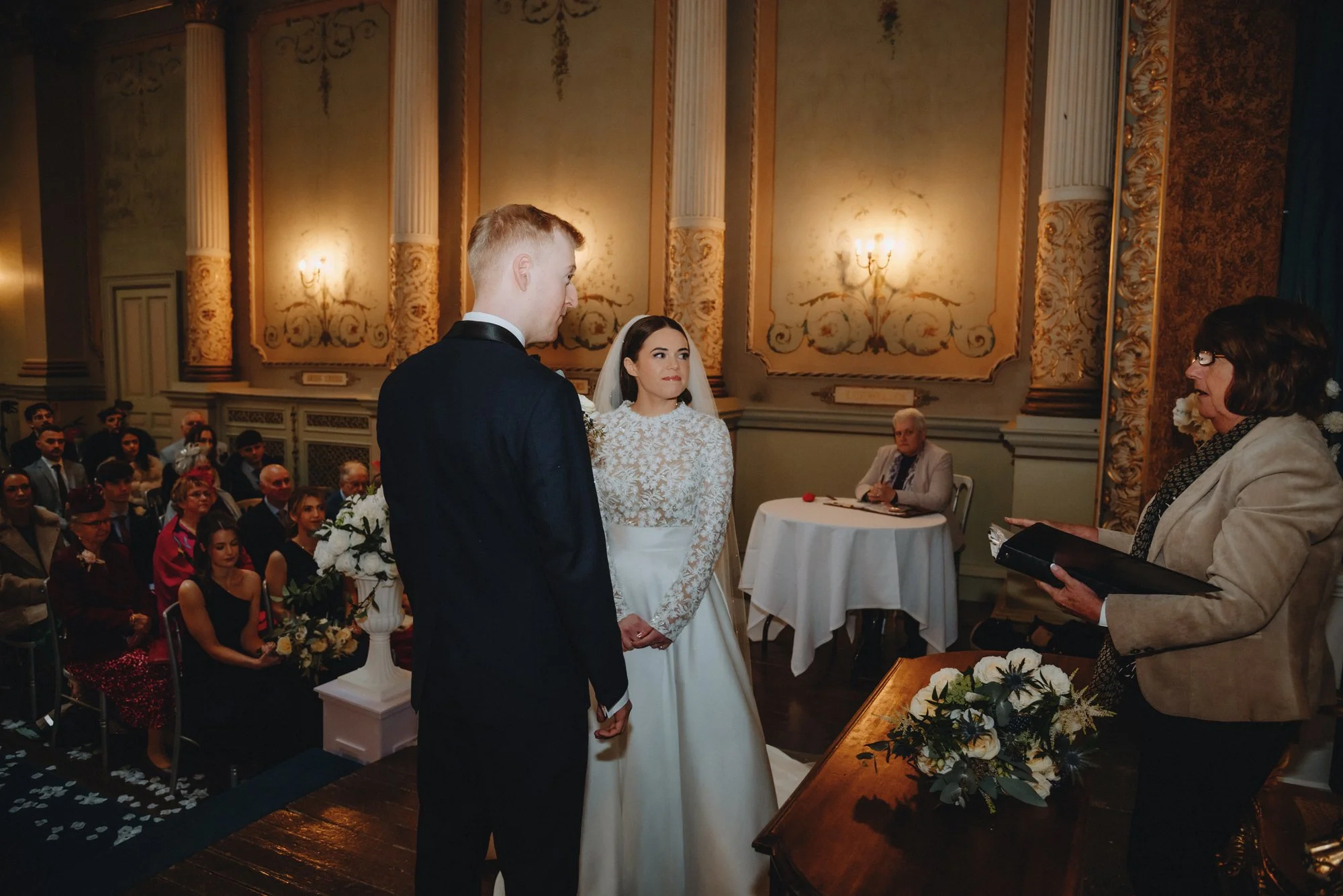A wedding ceremony with a bride and groom standing in front of an officiant. The bride is wearing a white lace dress and veil, the groom is in a dark suit. Guests are seated in the background inside a decorated ornate room.