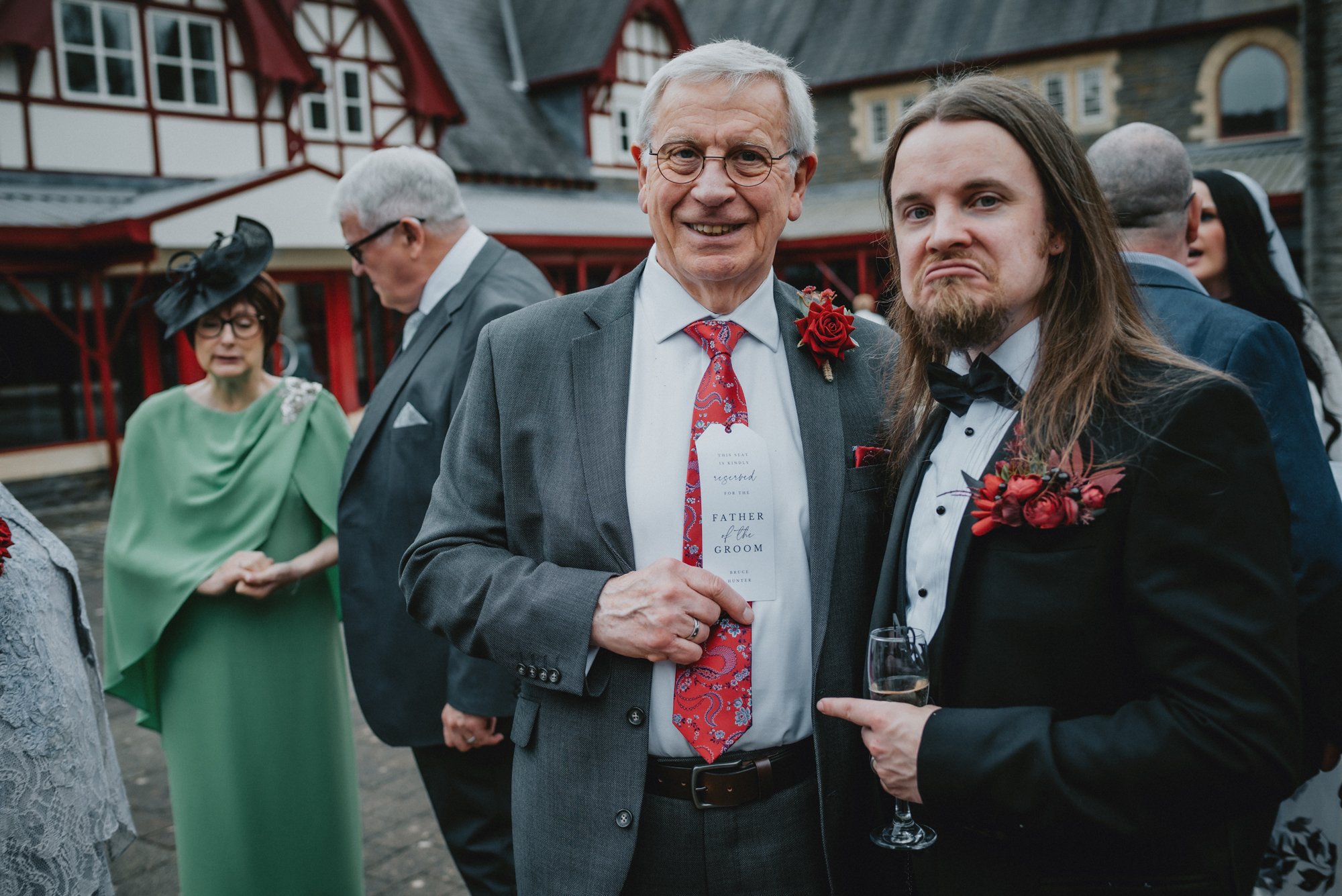 At a wedding reception outdoors, two men in suits are smiling and posing for the camera. One is an older man with glasses, a gray suit, and a red floral boutonniere, holding a reservation card that reads 'Father of the Groom.' The other is a younger 