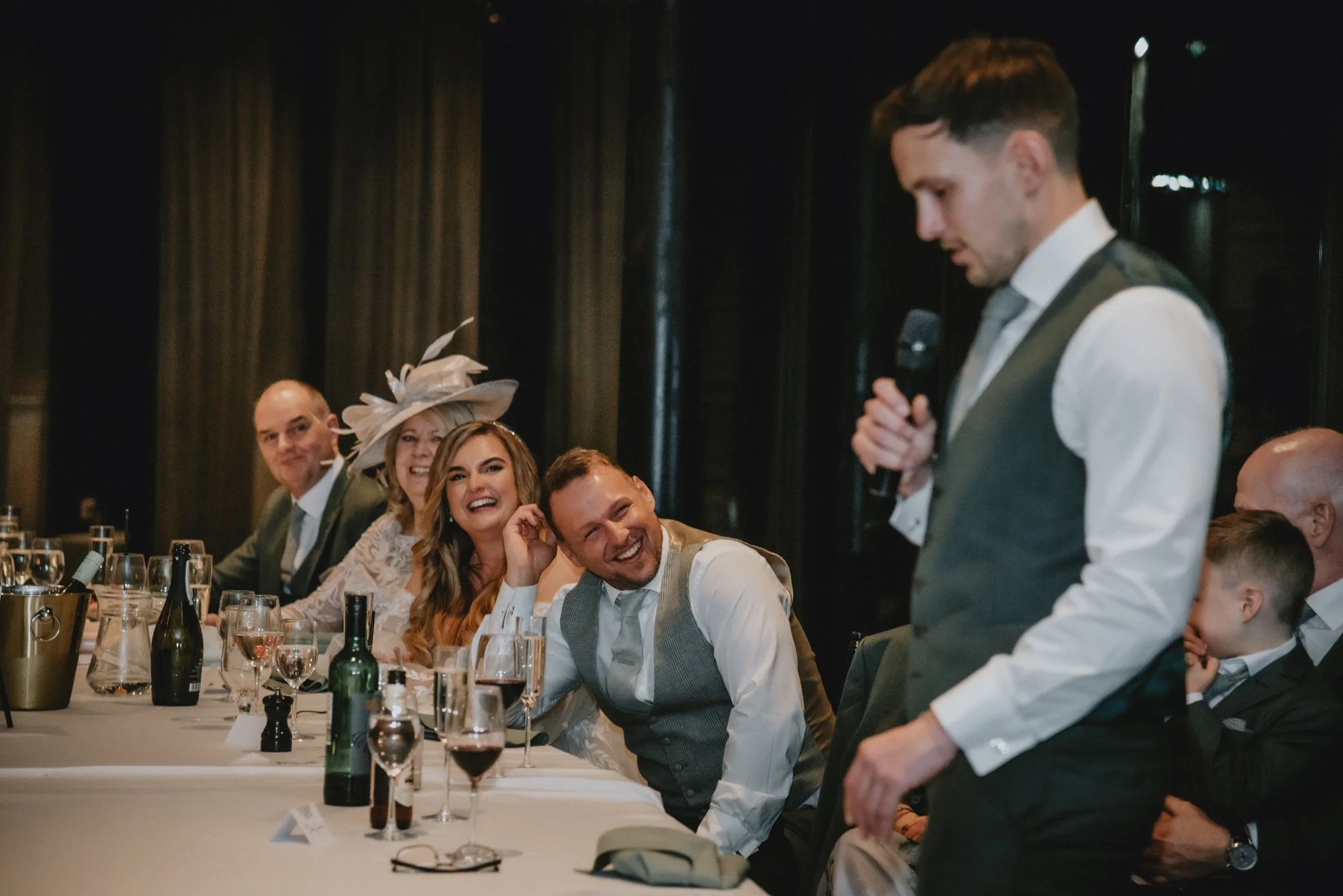 Group of people seated at a formal event, listening to a man giving a speech while holding a microphone; they are dressed in formal attire, with some smiling and laughing.