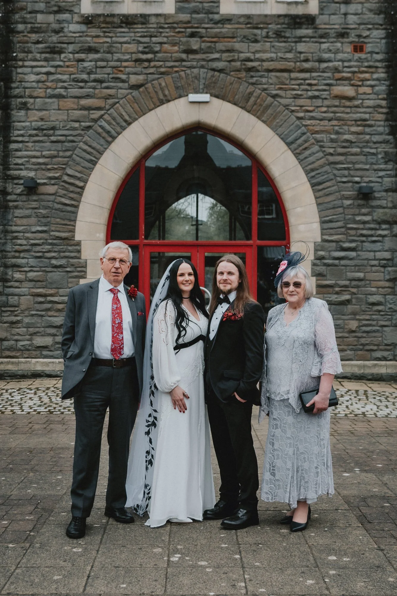 A wedding party standing in front of a brick building with a large arched window. The group includes a bride and groom, two older adults, and each person dressed in formal attire.