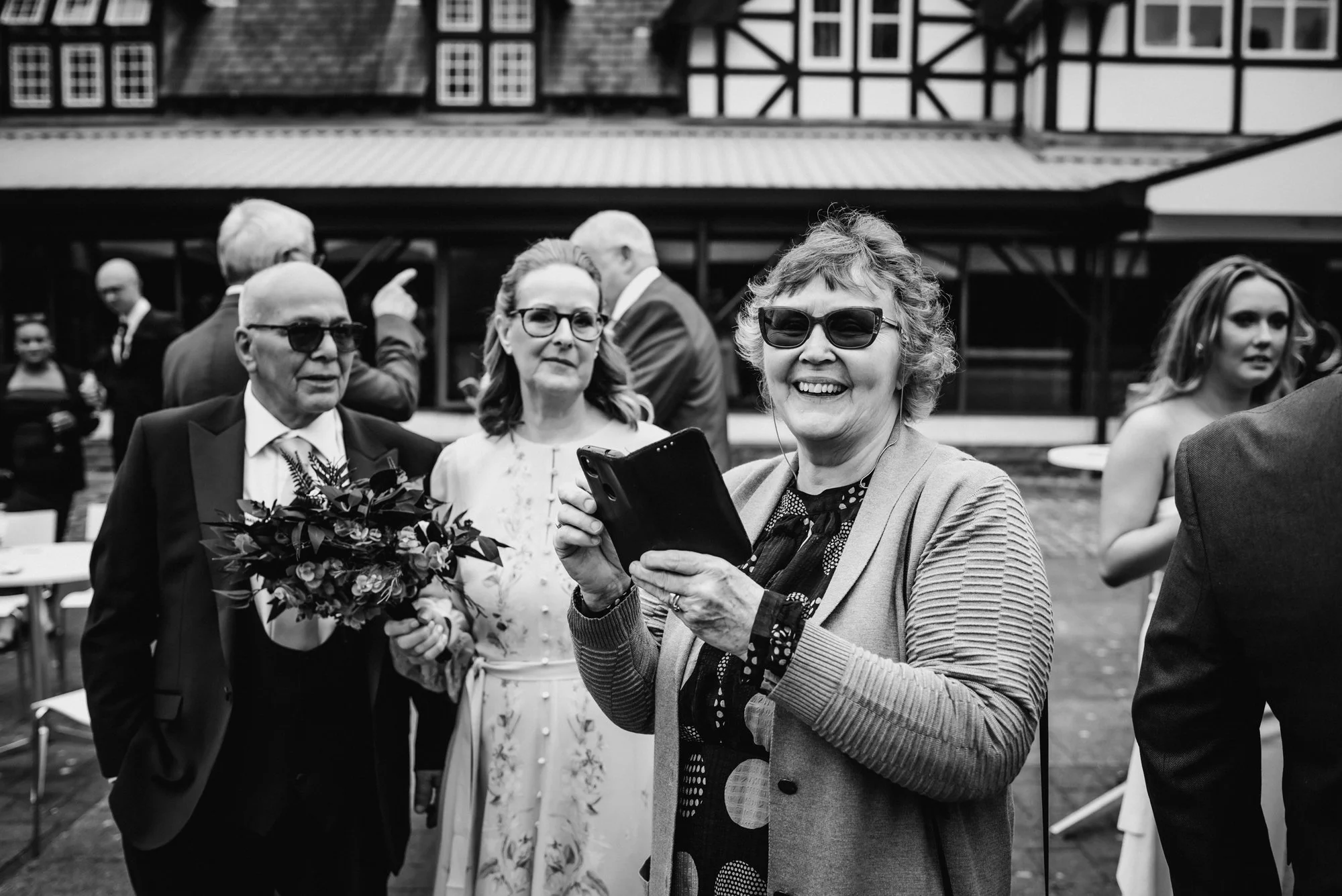 A group of people at an outdoor event, including a smiling woman in sunglasses holding a smartphone, a man in sunglasses holding a bouquet of flowers, and two women in the background, with a building with timber framing behind them.