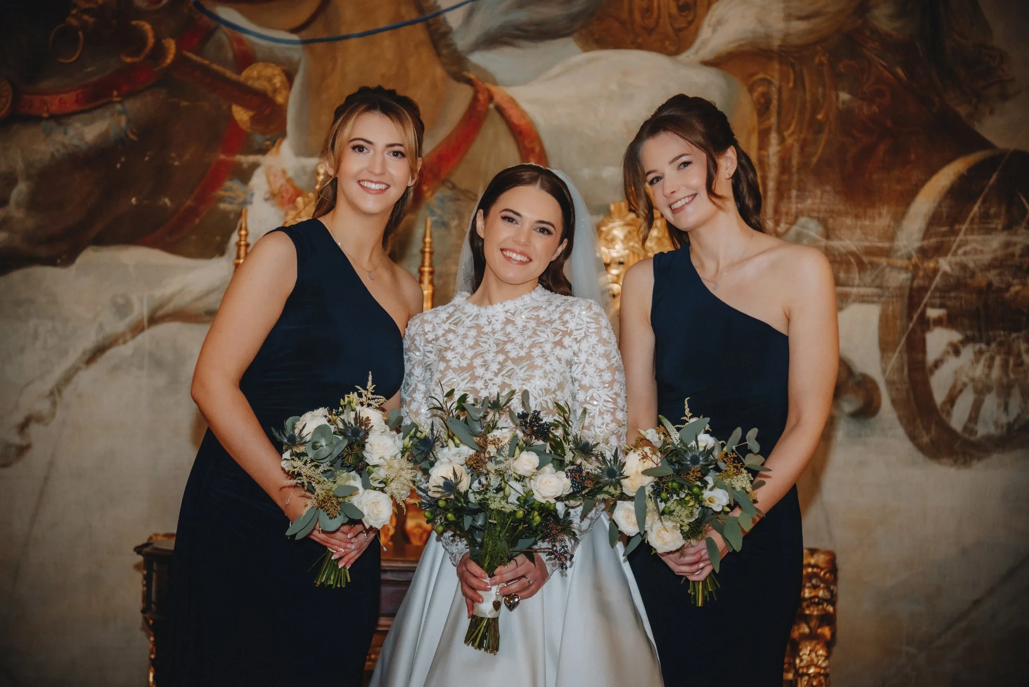 A bride in a white lace wedding dress and veil stands in the center, holding a bouquet of white and green flowers, flanked by two women in navy blue dresses holding matching bouquets, in front of a decorative mural backdrop.