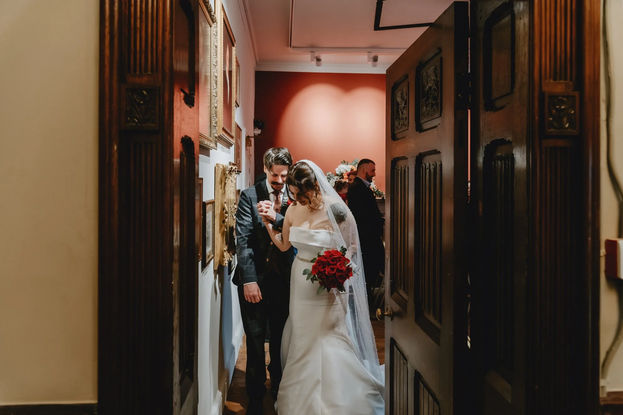 A bride and groom standing close together, holding hands with heads bowed, in a cozy indoor setting with framed pictures on the wall, a woman in a dark dress and other guests in the background.