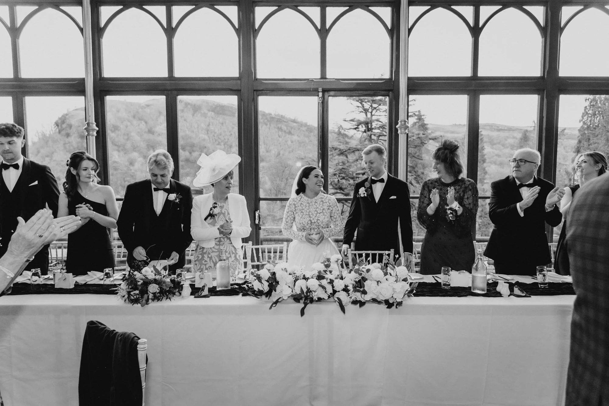 A wedding reception with the bride and groom standing at the table, surrounded by wedding guests clapping and smiling in front of a large window with scenic outdoor view.