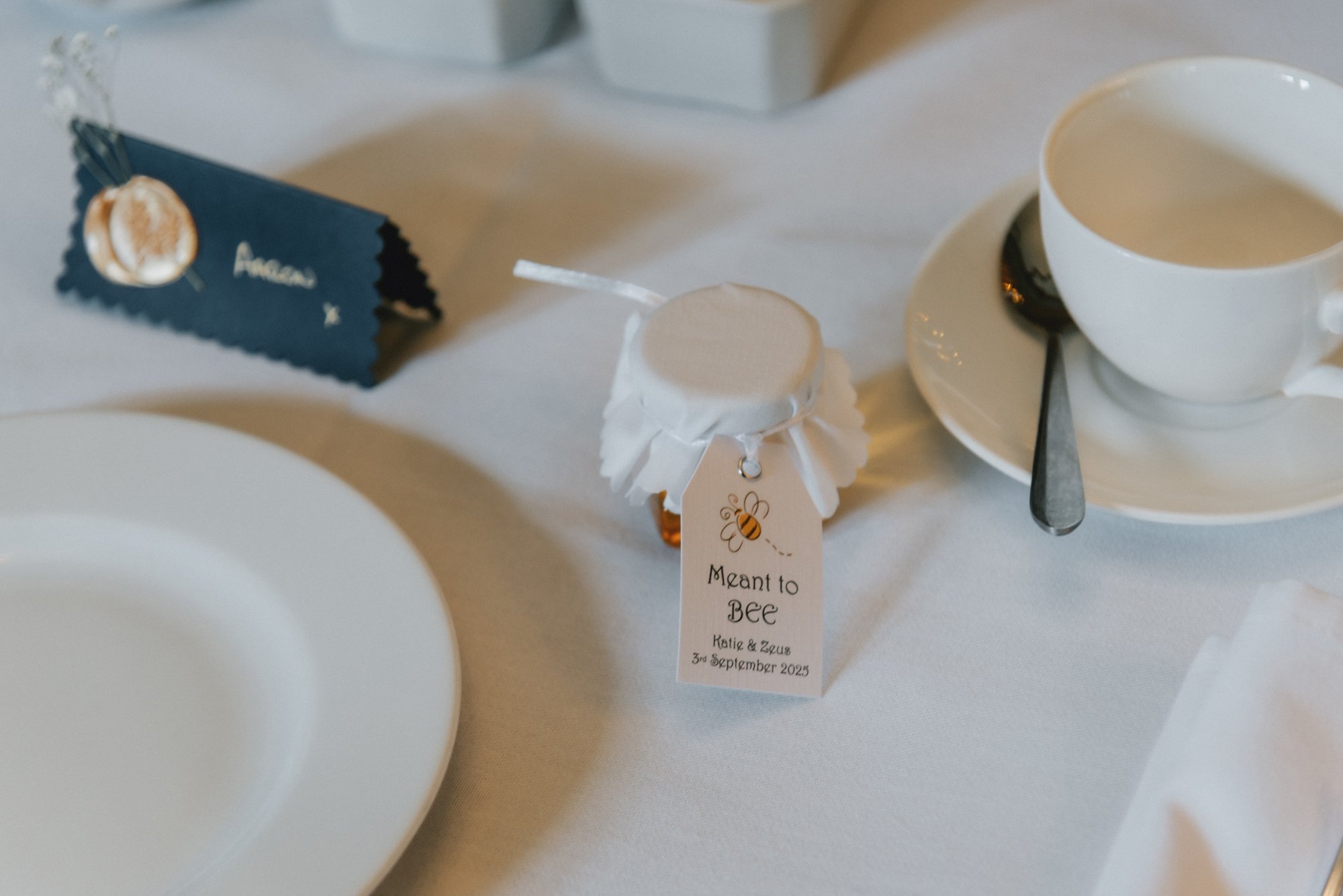 A wedding table setting featuring a white tablecloth, a small jar with a bee and flower illustration and a tag that reads 'Meant to BEE Katie & Zeus 3rd September 2025,' a white cup and saucer with a silver spoon, a white plate, and a blue place card