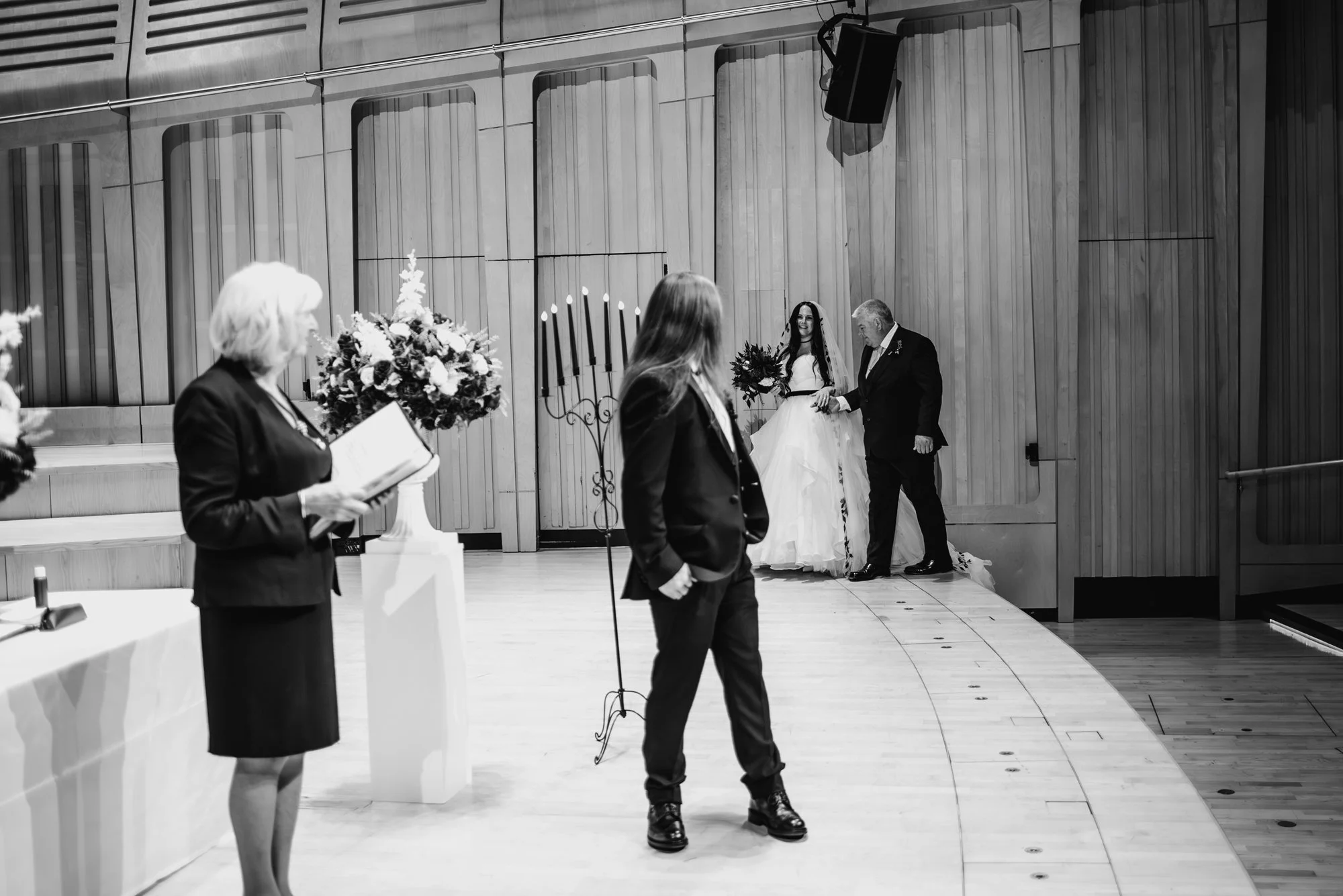 Black and white photo of a wedding ceremony with the bride and an older man, possibly her father, walking down the aisle. A woman is reading from a book at a table, and a person in a suit stands in the foreground.