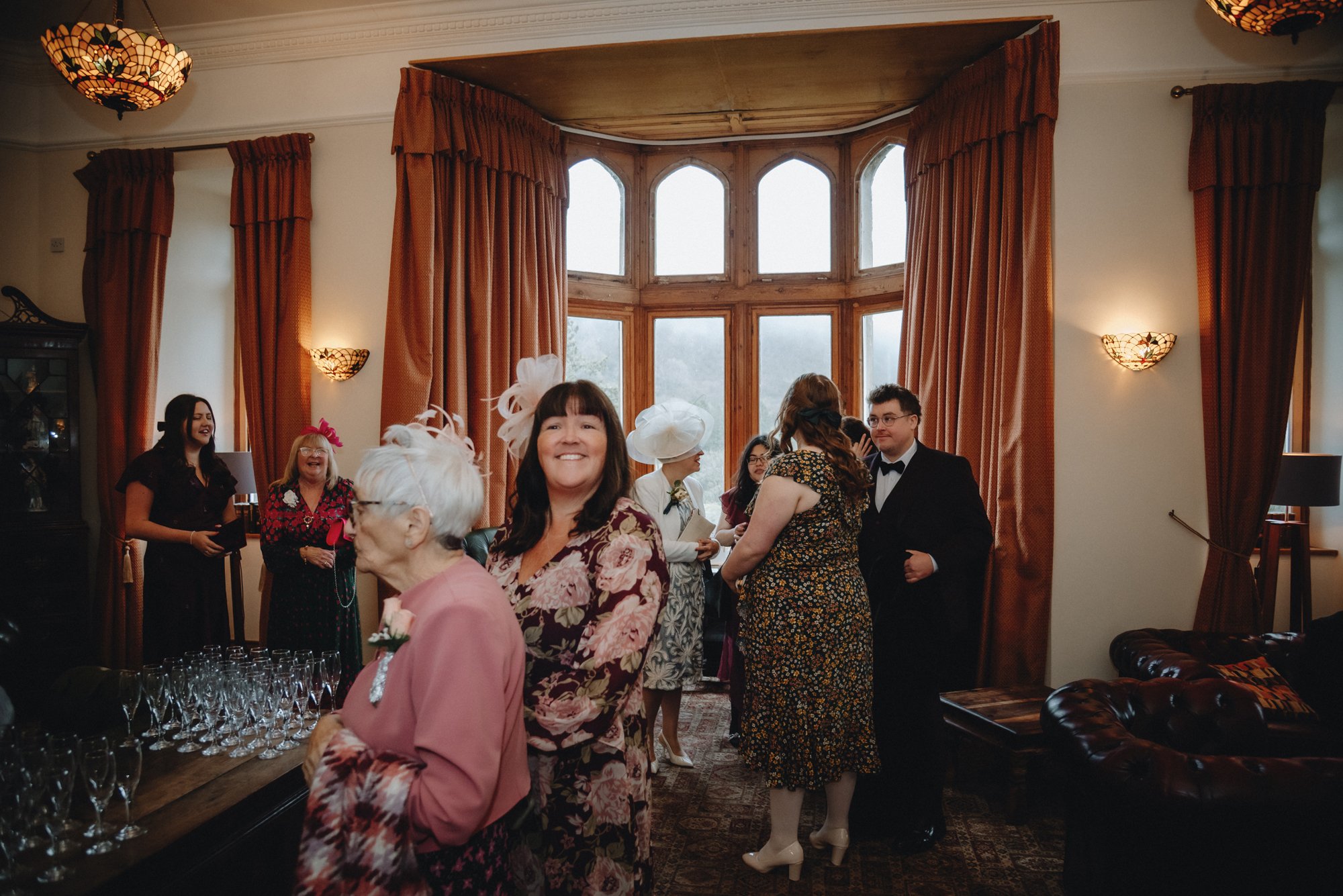 People gathering during a celebration in a room with large bay windows, orange curtains, and vintage decor. The group includes women with hats and a man in a tuxedo, chatting and smiling.