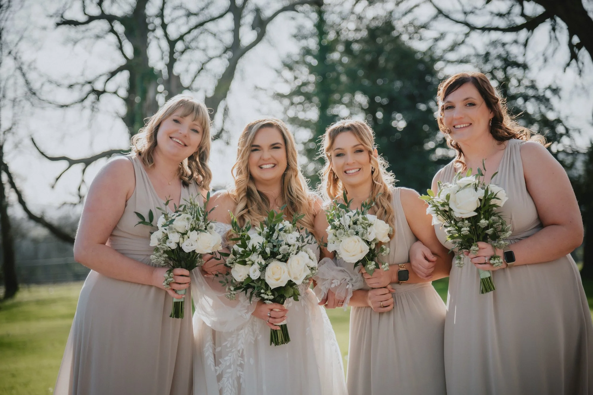 Four women in light-colored dresses holding bouquets of white flowers smiling outdoors on a sunny day.