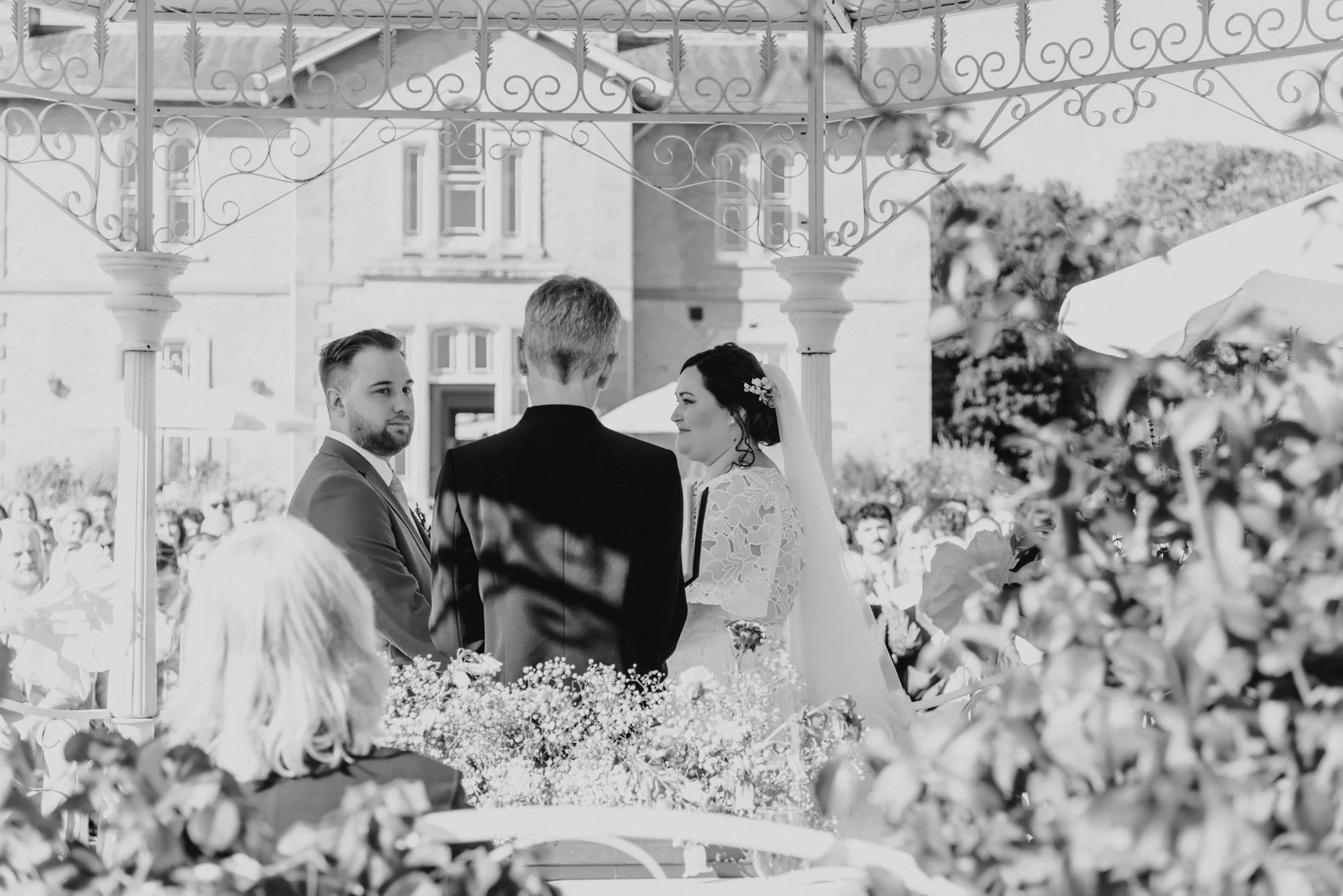 A black-and-white photo of a wedding ceremony outdoors, with the bride and groom facing each other and a male officiant standing between them. The bride is wearing a lace dress and veil, and the groom in a suit. There are flowers in the foreground, a