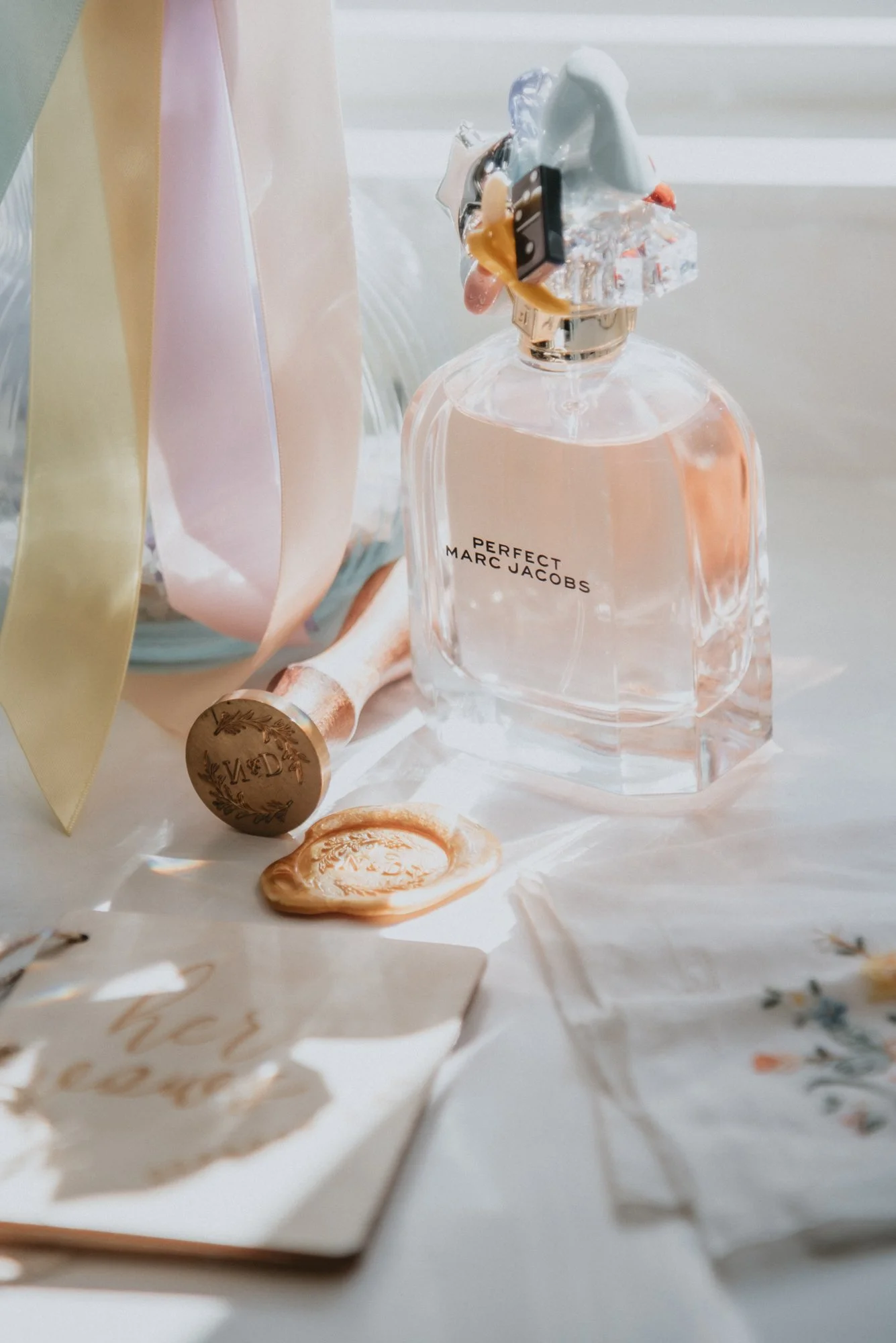 A perfume bottle labeled 'Perfect Marc Jacobs' with a decorative cap, surrounded by a wax seal, a handwritten note, and colorful ribbons on a white surface in natural light.