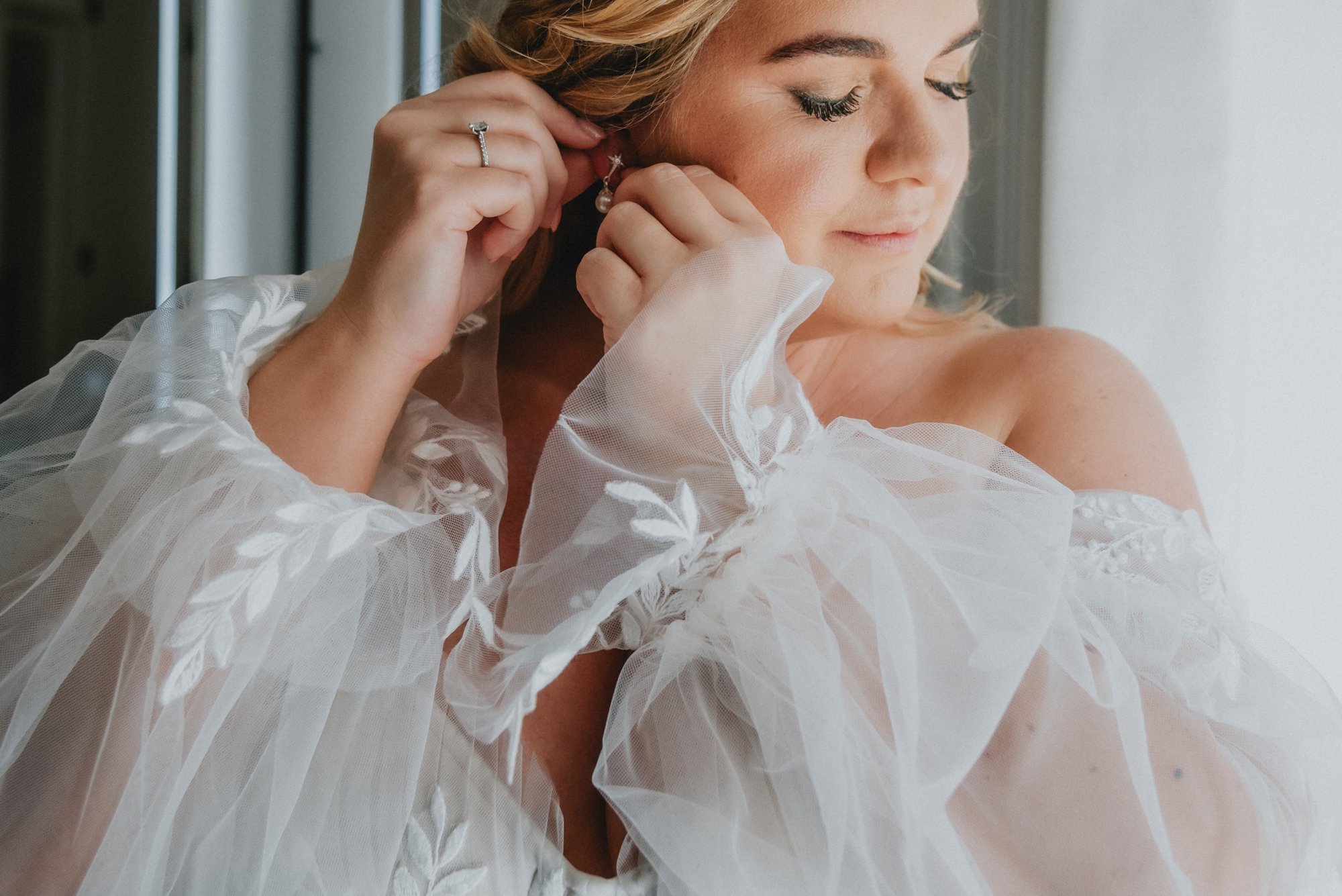 A woman dressing up, adjusting earrings, wearing a wedding dress with puffy sheer sleeves, near a window with light coming in.