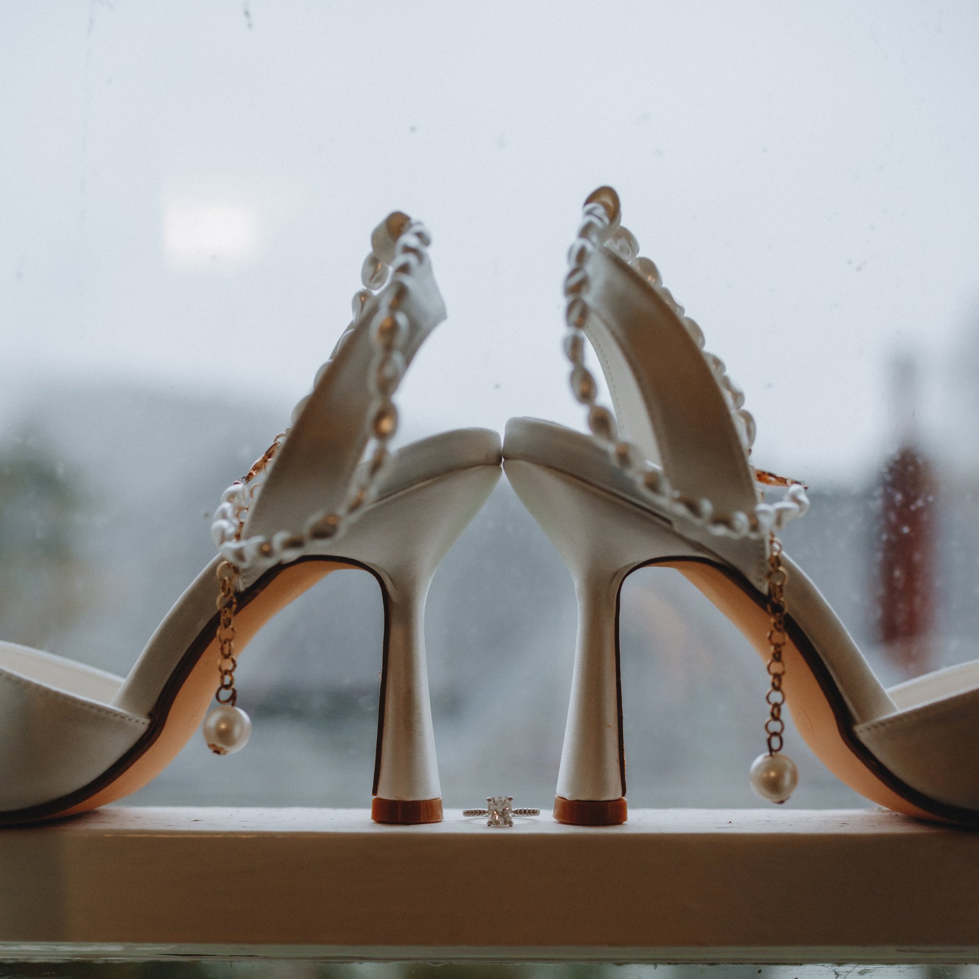 A pair of white high-heeled shoes decorated with pearl accents, upside down with heels touching, with an engagement ring placed between the heels, against a rainy window background.